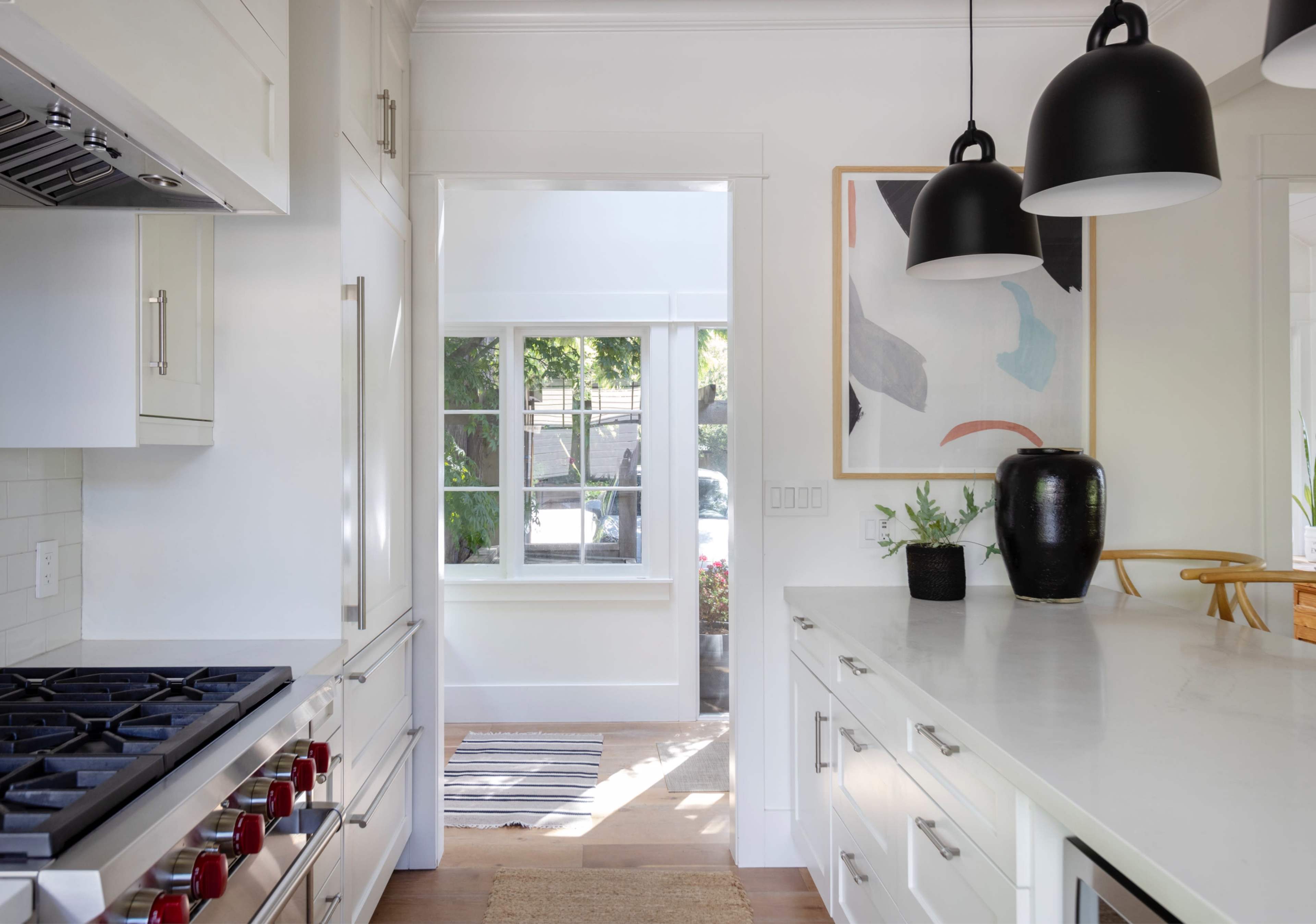 A modern kitchen with white cabinetry, a large gas stove, and a view of a hallway leading to a well-lit area with an abstract painting and plants.