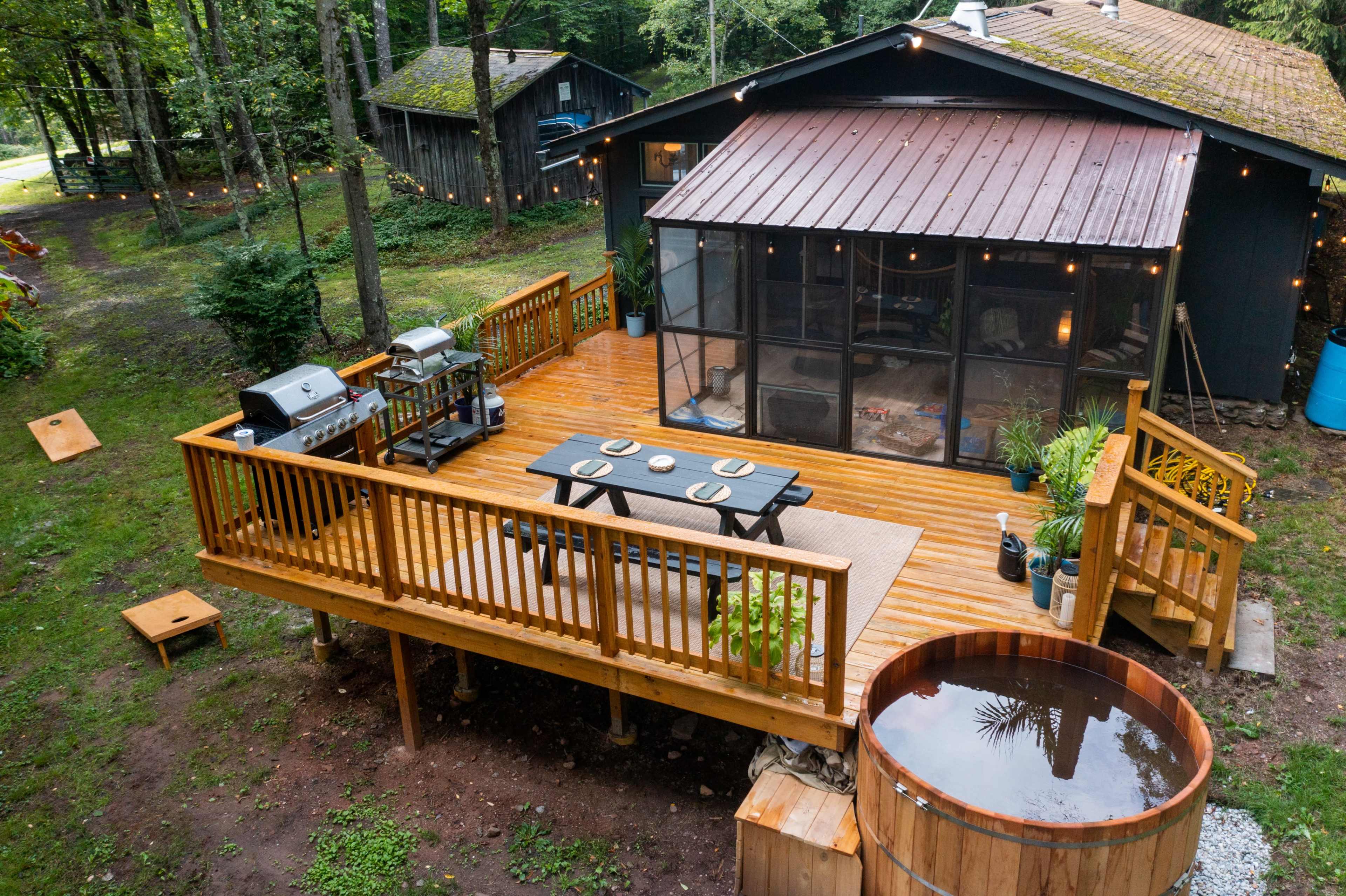 A wooden deck with a dining table and chairs is attached to a house, featuring a hot tub and grill, surrounded by trees and outdoor seating.
