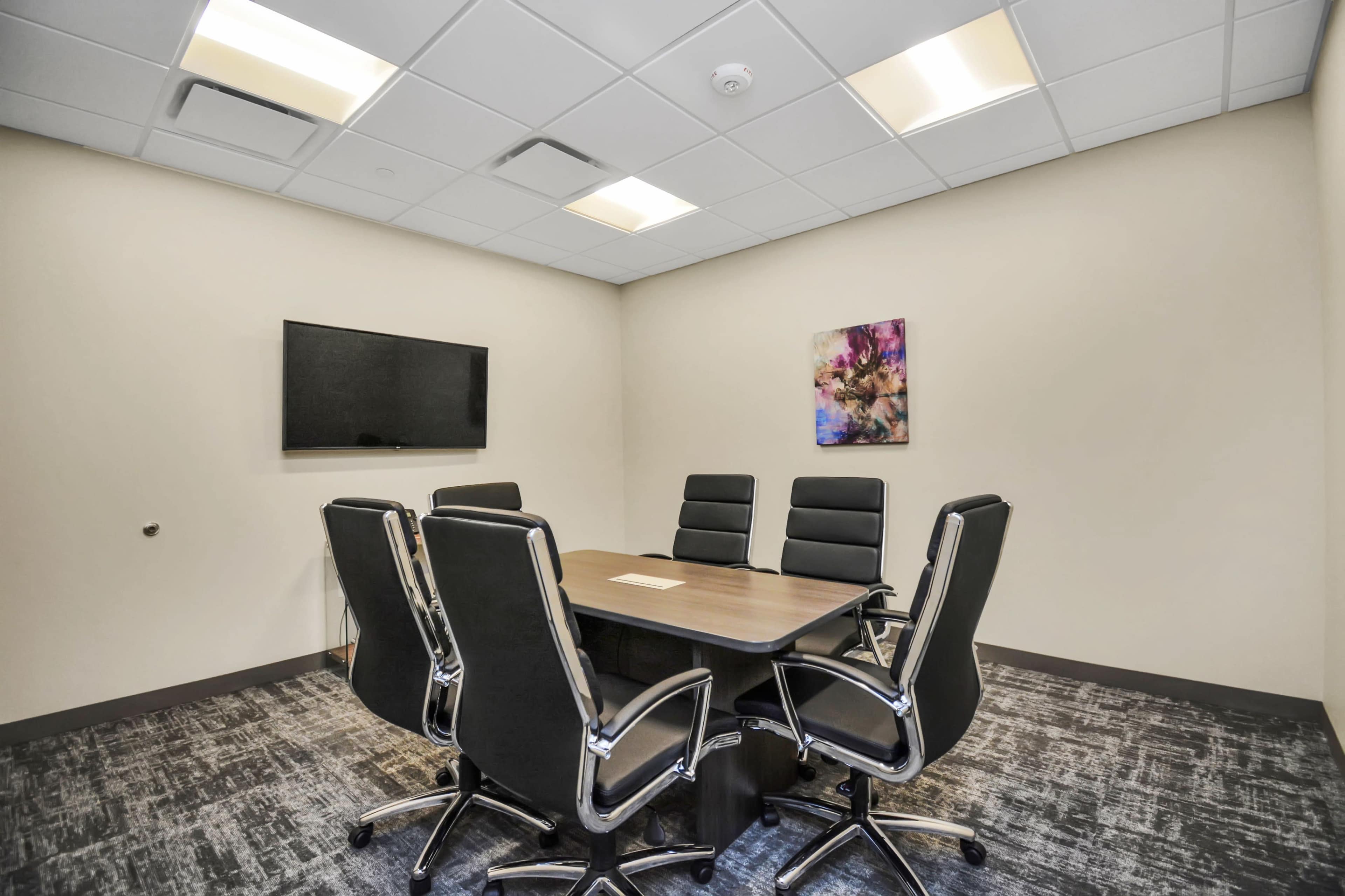 A modern conference room features a large wooden table surrounded by six black leather chairs and a wall-mounted television.