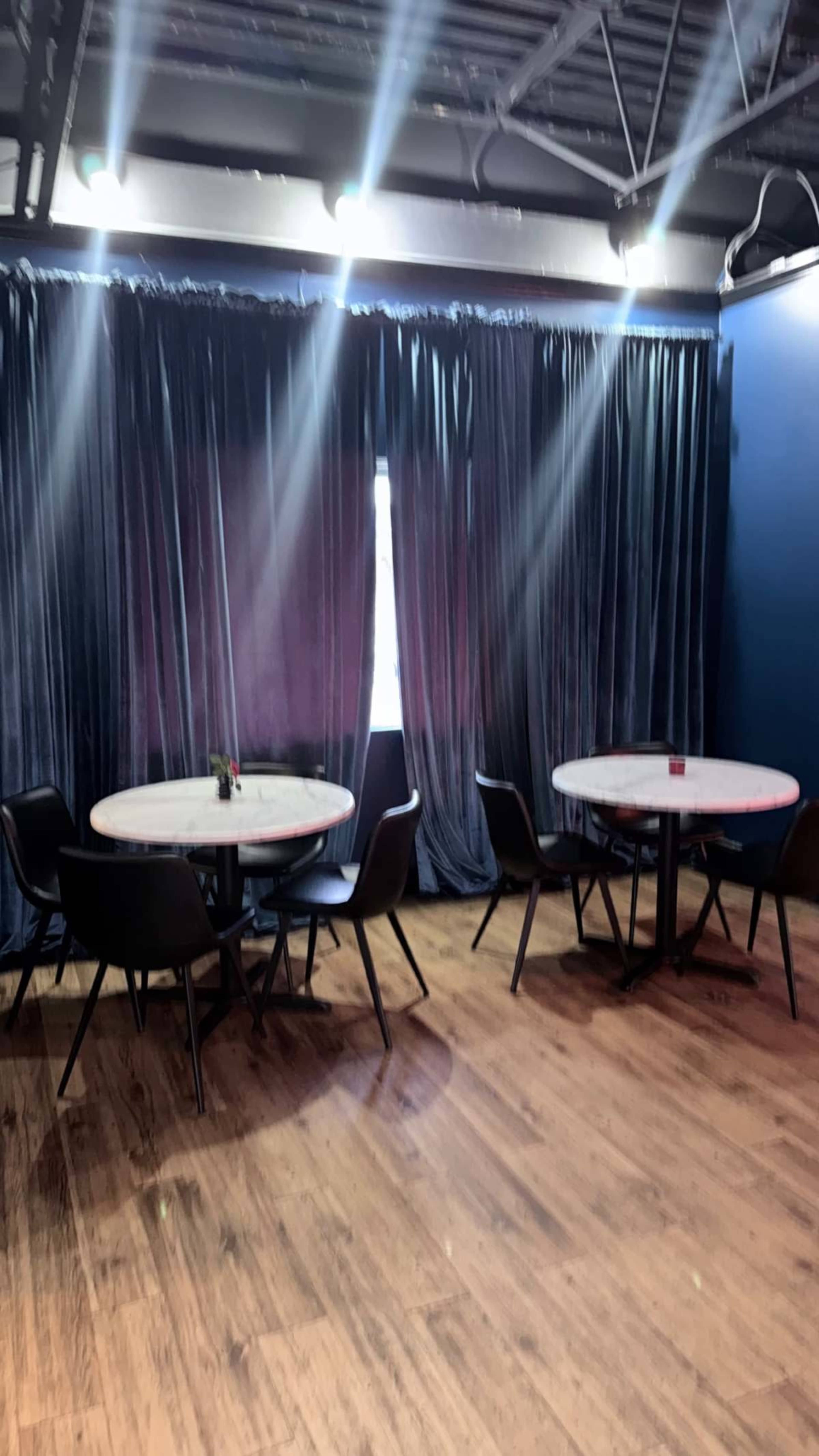 The image shows a modern café space with two round tables surrounded by black chairs, illuminated by soft overhead lighting and framed by dark curtains.