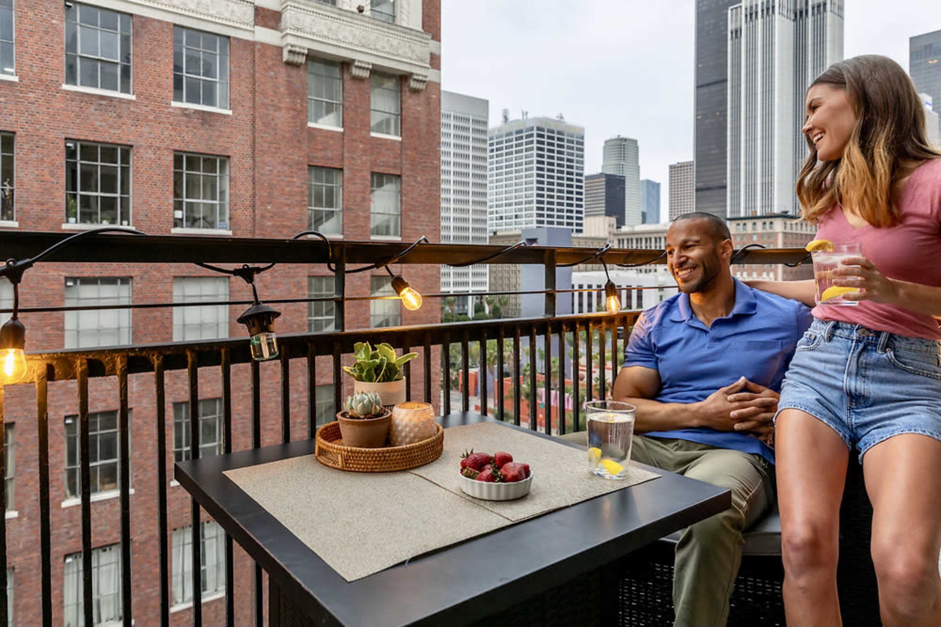 A man and woman sit on a balcony with a view of a cityscape, enjoying drinks and fresh strawberries on a table adorned with plants and fairy lights.