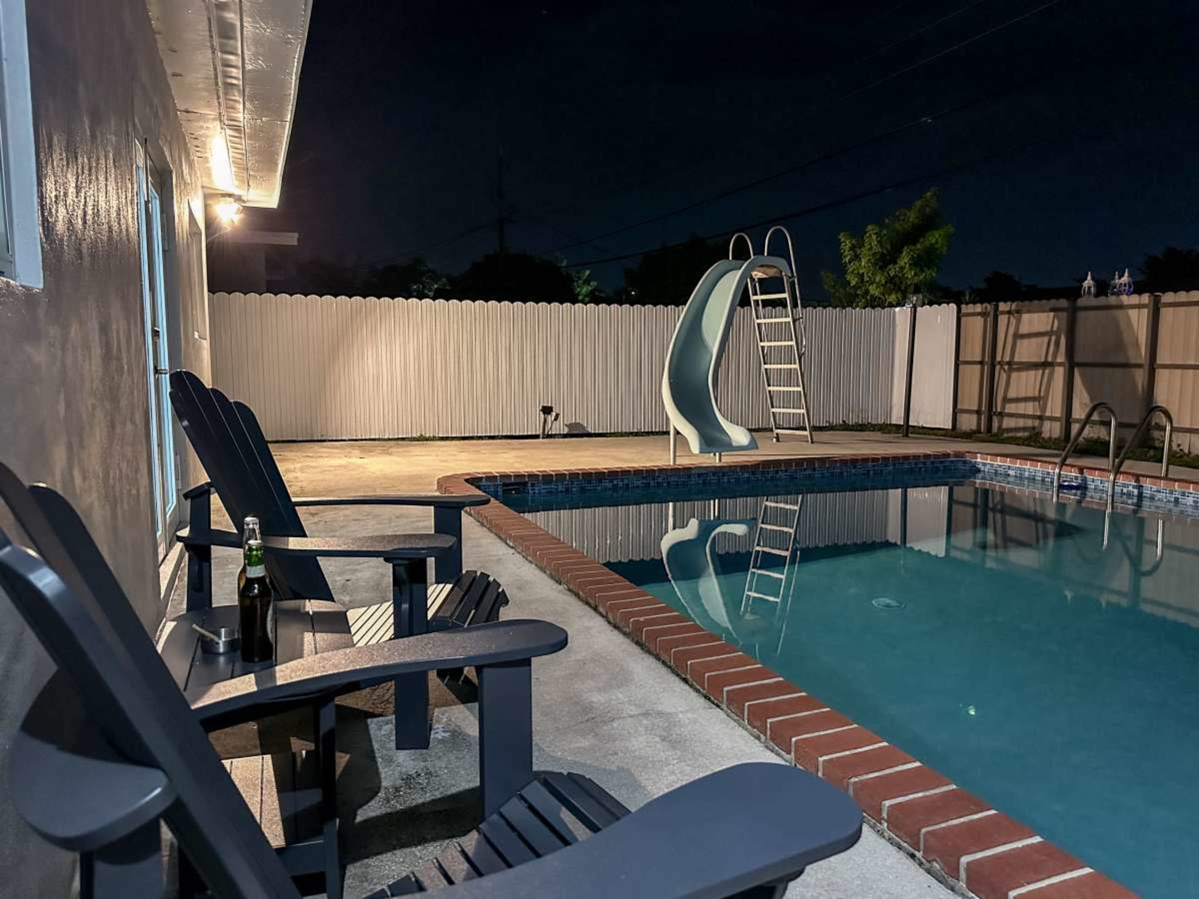 The image shows a nighttime view of a swimming pool area with two chairs, a water slide, and a ladder, surrounded by a white fence.