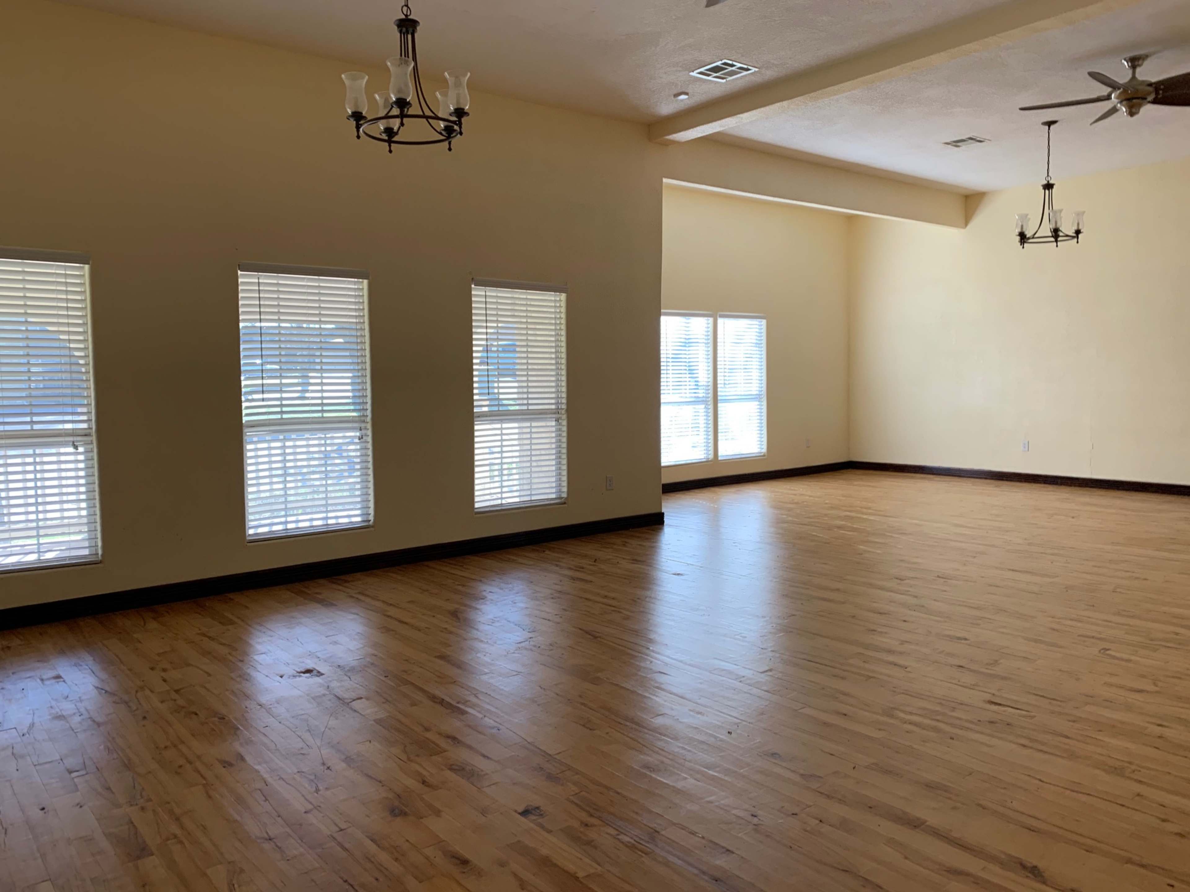 A spacious room with hardwood flooring, four large windows featuring blinds, and two ceiling fans.