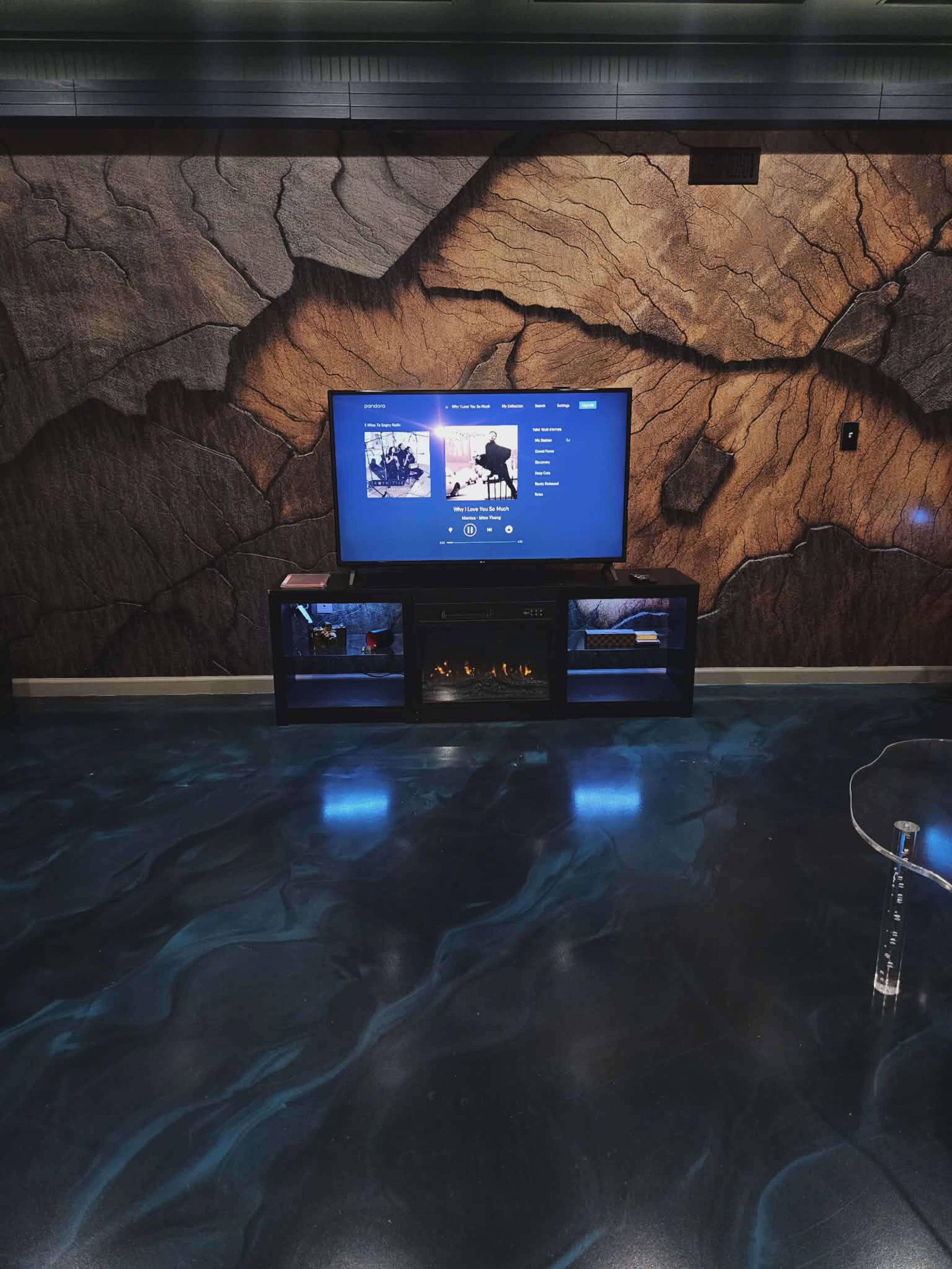 A television mounted on a wall displays a media interface against a textured, rocky backdrop, with an entertainment center featuring an electric fireplace below it.