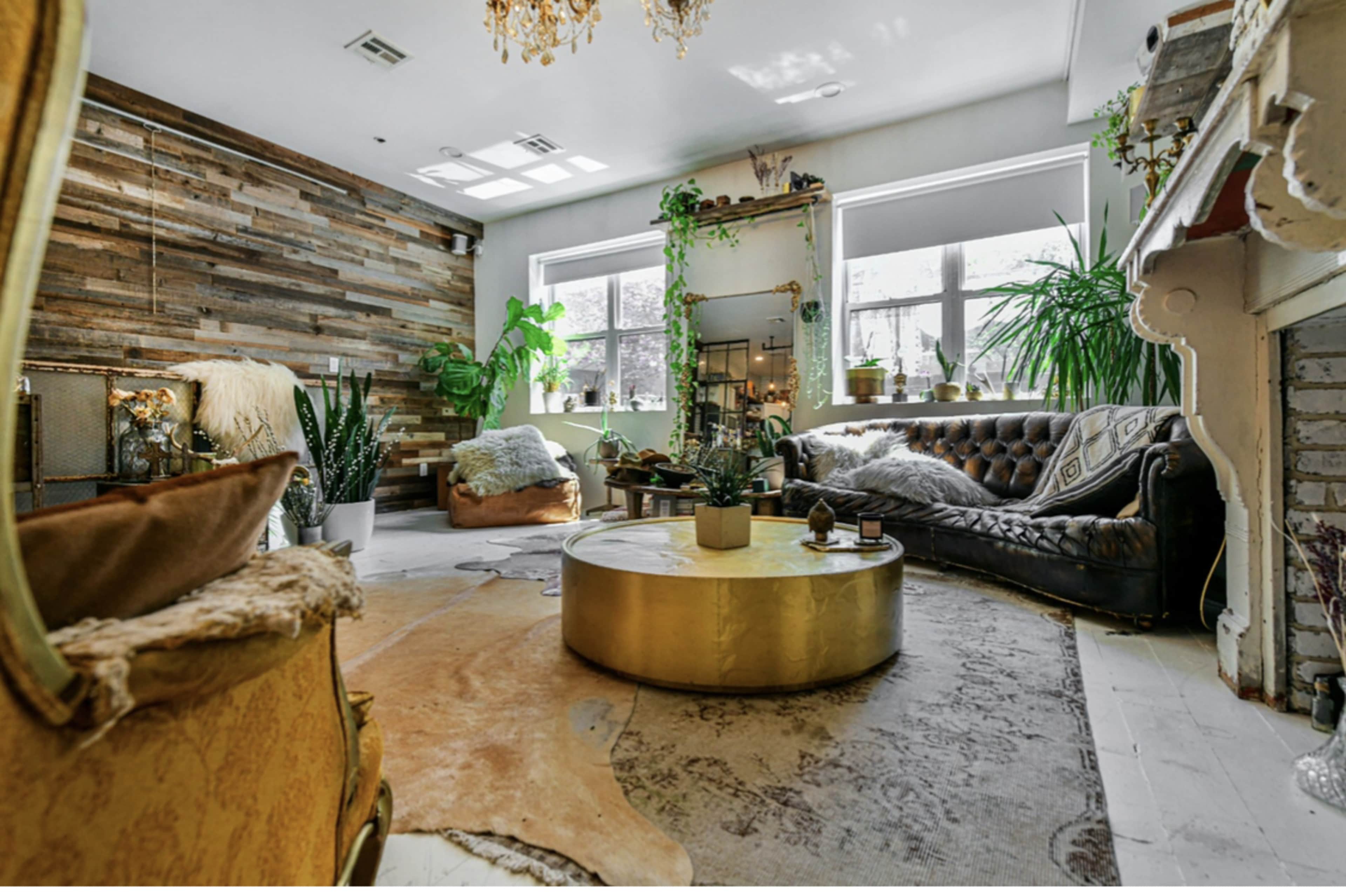 The image shows a cozy living room with a mix of vintage and modern decor, featuring a leather sofa, a circular gold coffee table, and large windows surrounded by indoor plants.