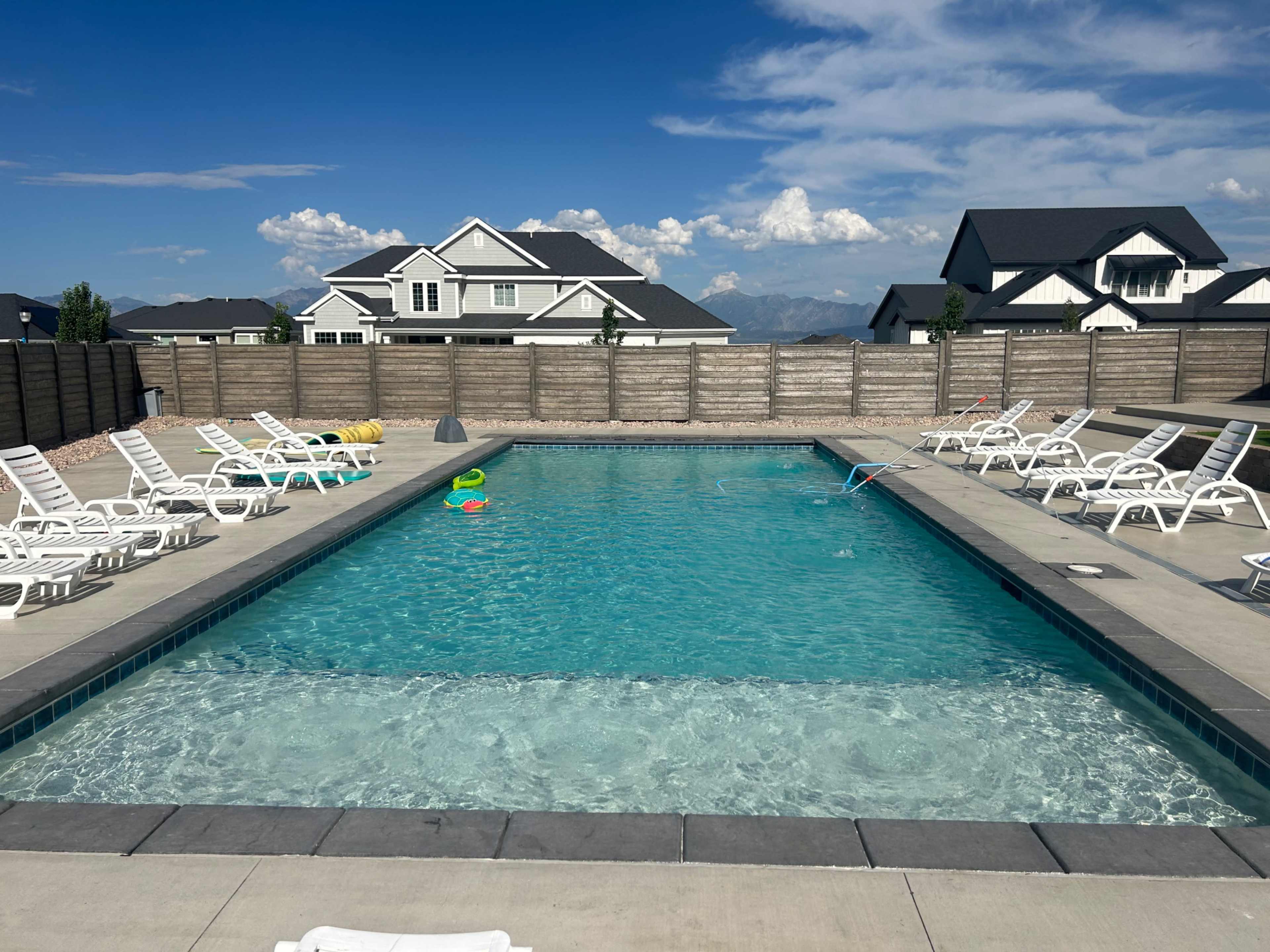 A clear swimming pool surrounded by lounge chairs, with a wooden fence in the background and houses beyond it.
