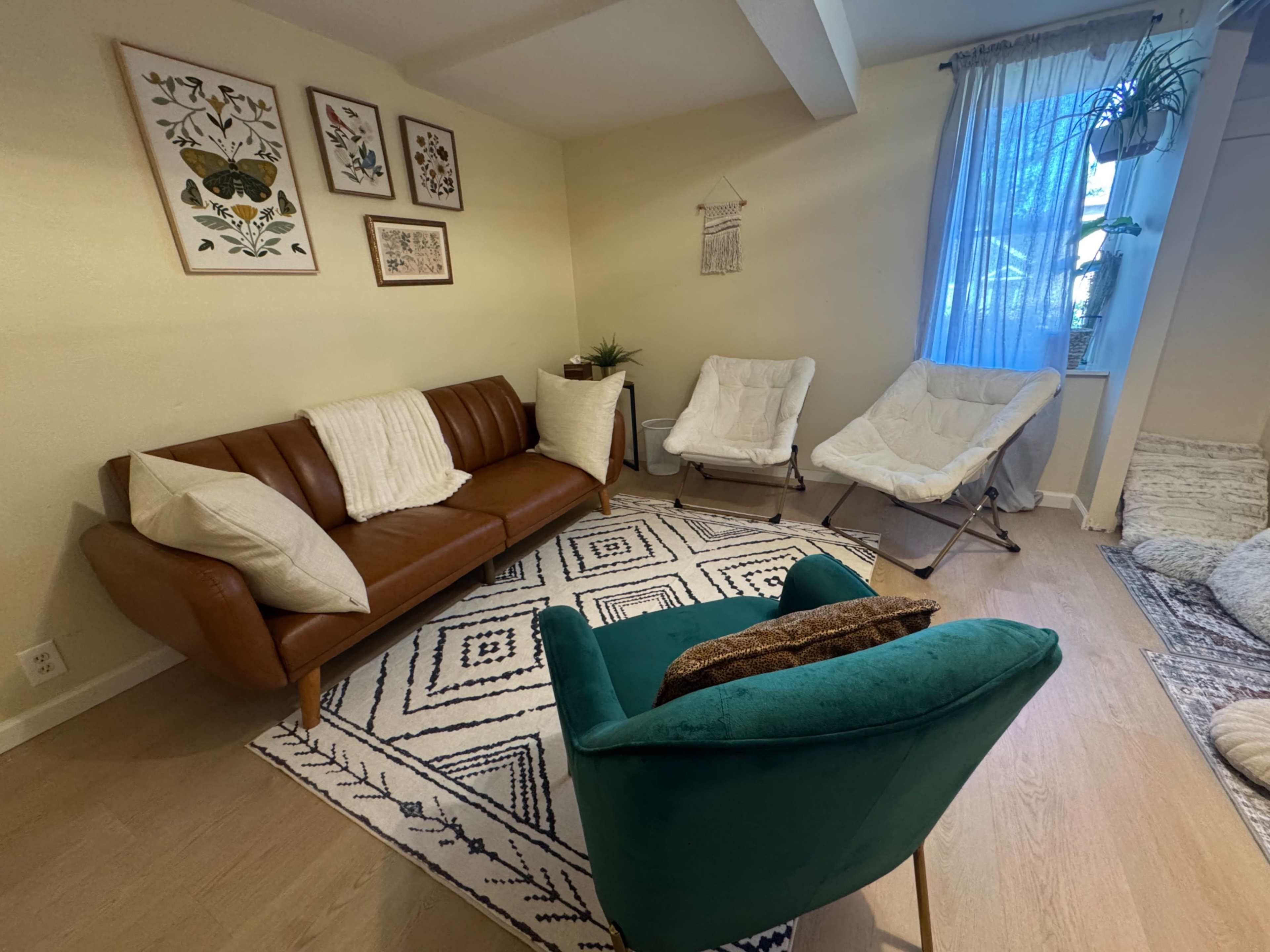 The image shows a cozy living room with a brown leather sofa, two white lounge chairs, a green accent chair, and decorative wall art, all arranged around a patterned area rug.