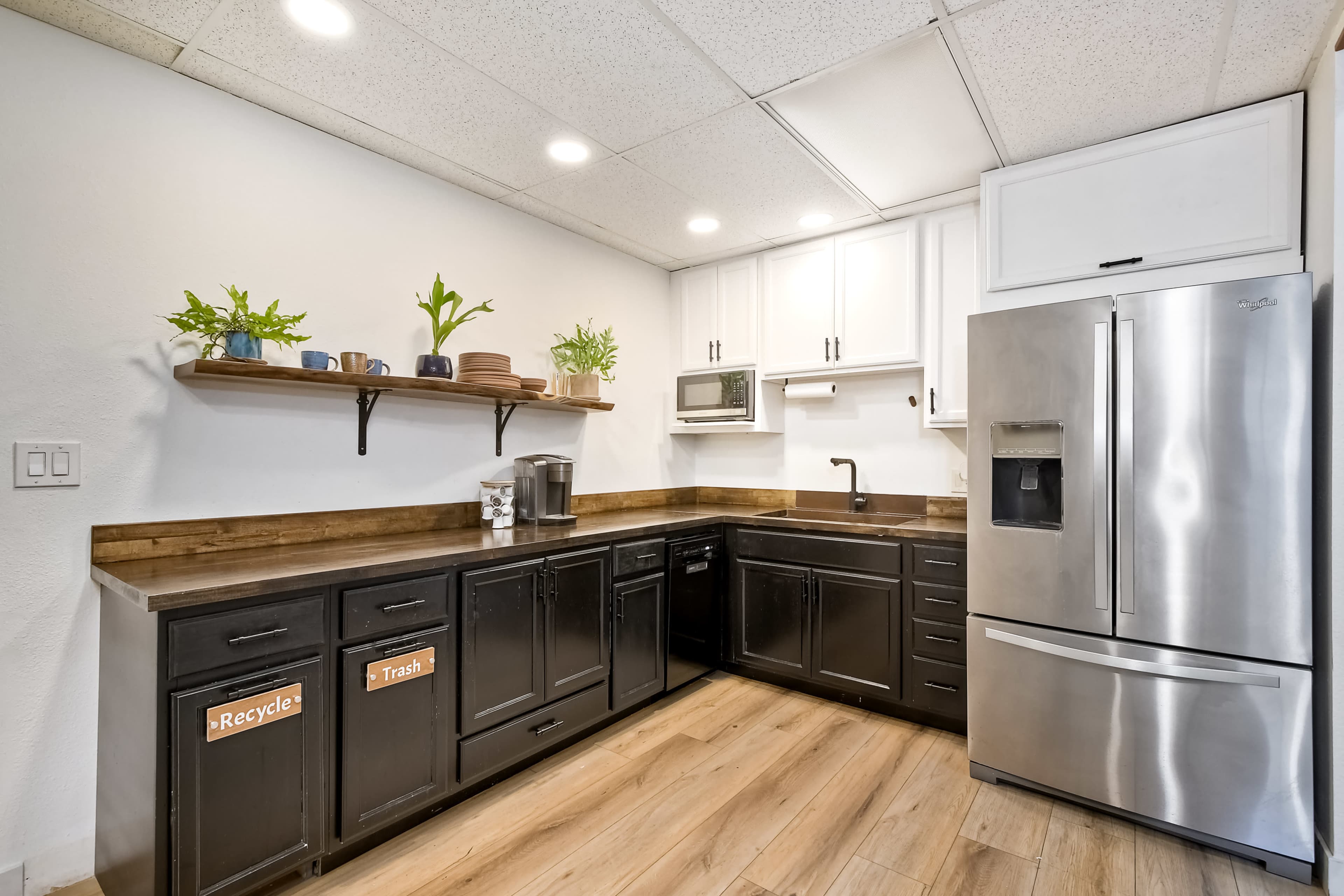 The kitchen features a dark cabinetry with labeled recycling and trash bins, a stainless steel refrigerator, and simple shelving with decorative plants and dishware.