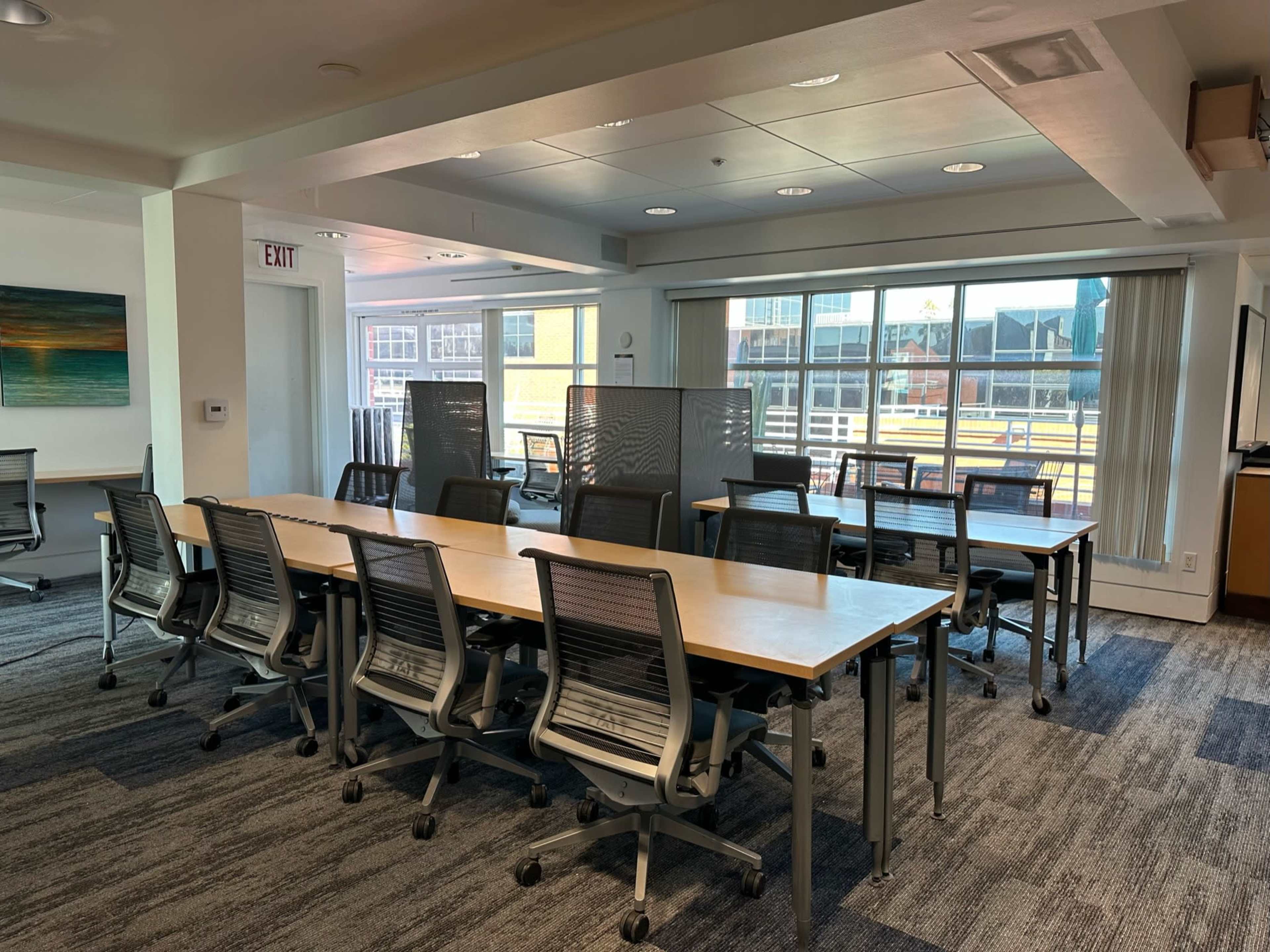 A modern office space with several rows of desks and ergonomic chairs arranged near large windows that allow natural light to enter.