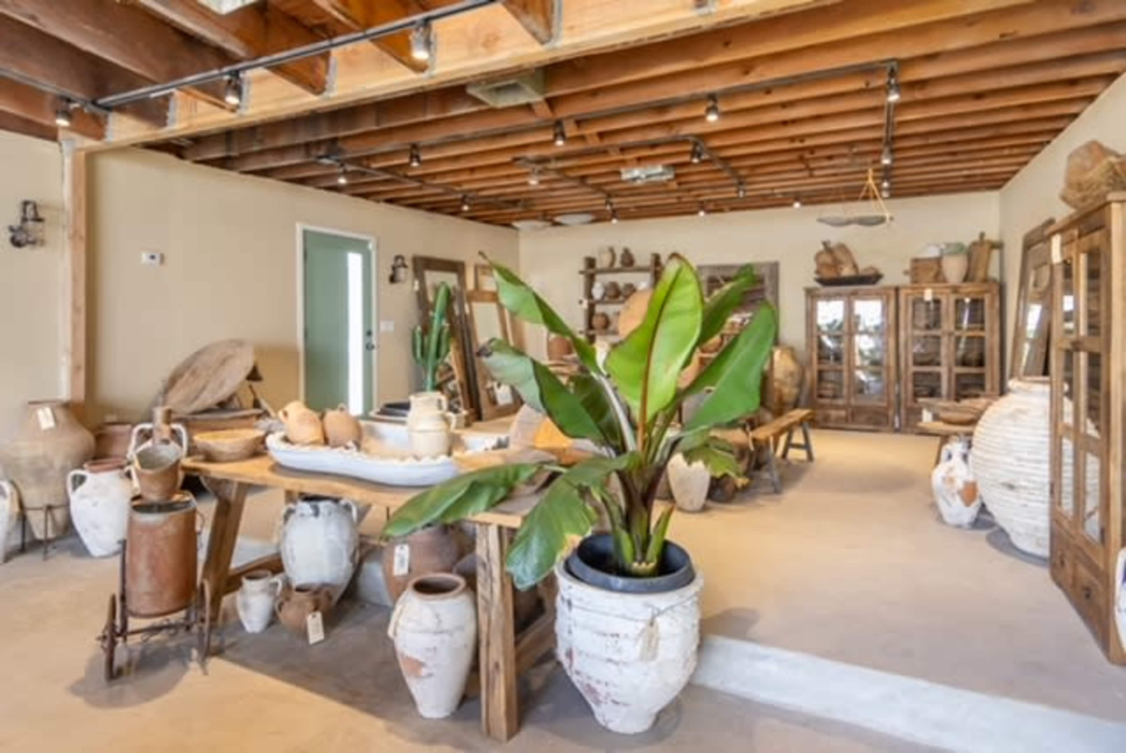 The image depicts a rustic interior space filled with various pottery and artifacts, arranged on tables and shelves beneath exposed wooden beams.