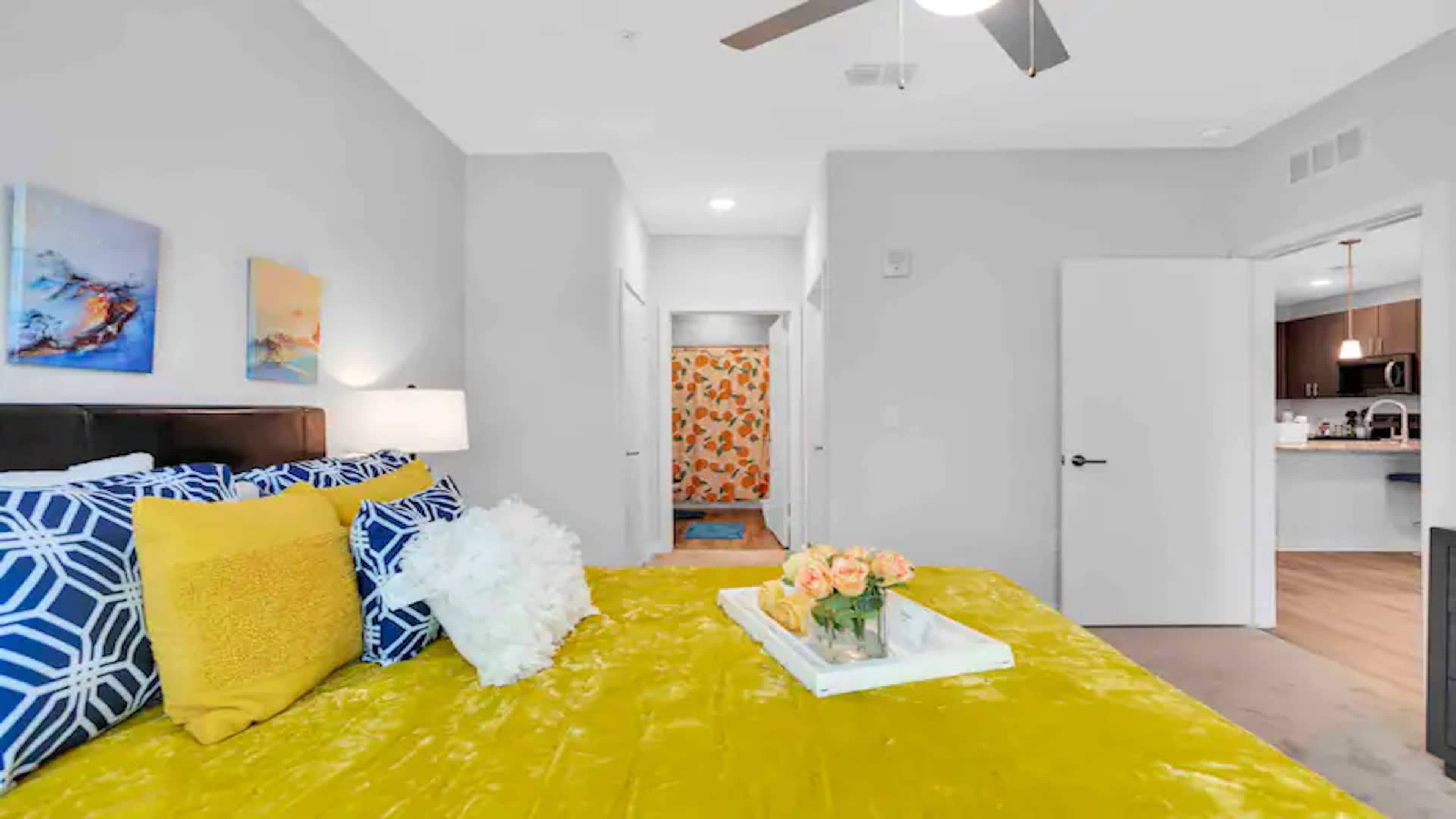The image shows a bedroom with a yellow bedspread, decorative pillows, and a tray of flowers on the bed, leading to a bathroom area with patterned curtains and a doorway to a kitchen.