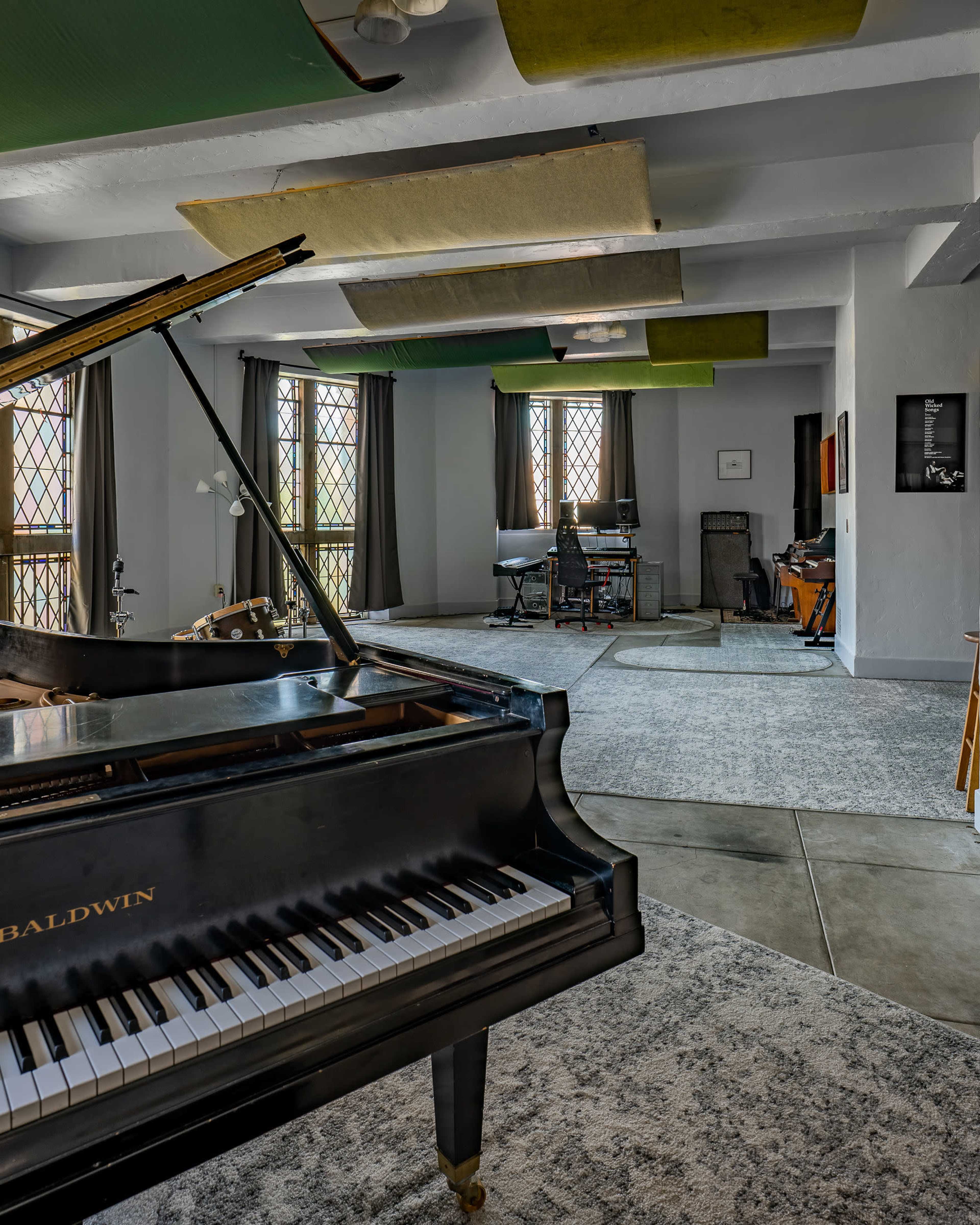 The image shows a spacious room featuring a grand piano in the foreground, with large windows and various musical instruments and furniture arranged throughout.
