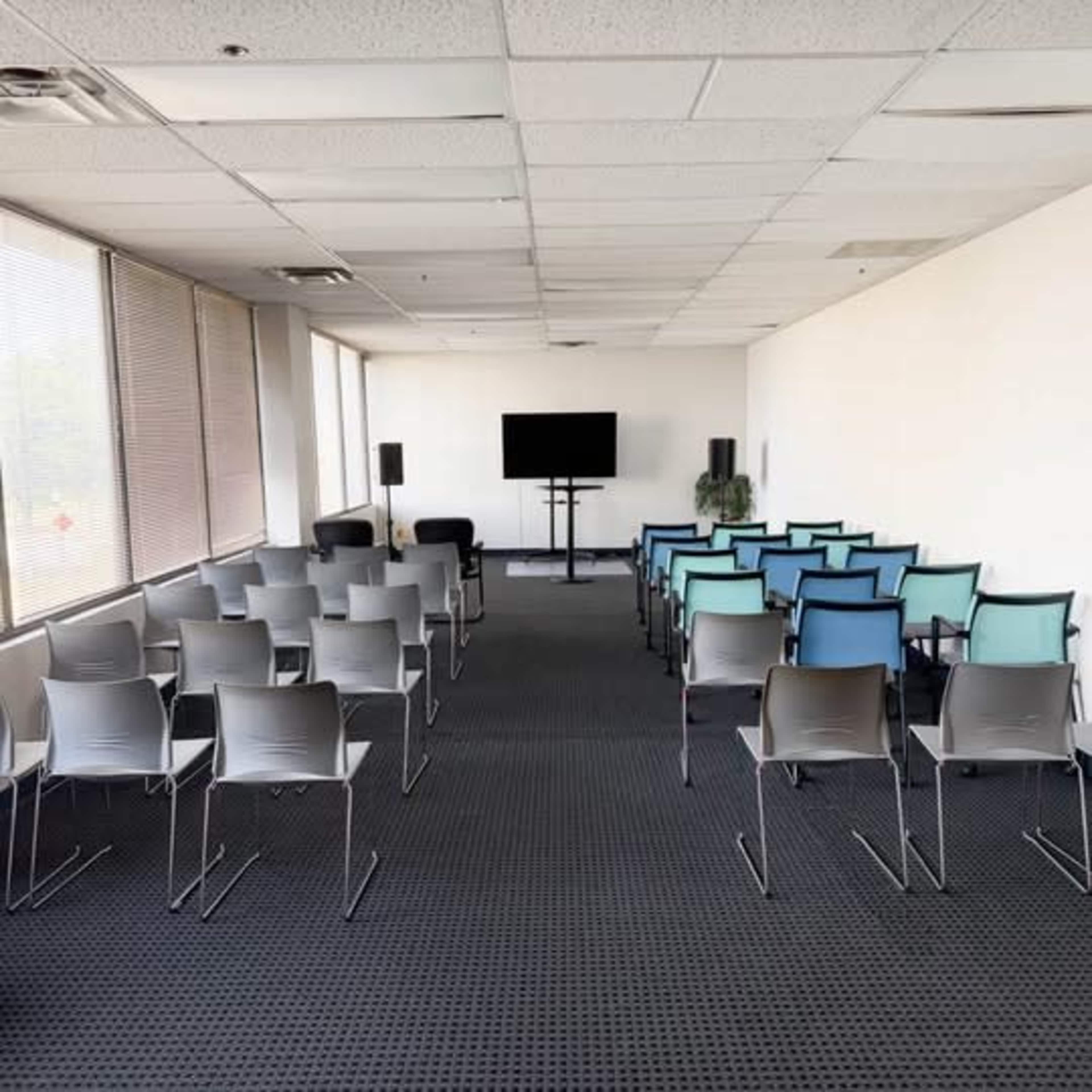 The image shows a spacious indoor room arranged with several rows of chairs facing a large screen and two microphones, with large windows on one side.