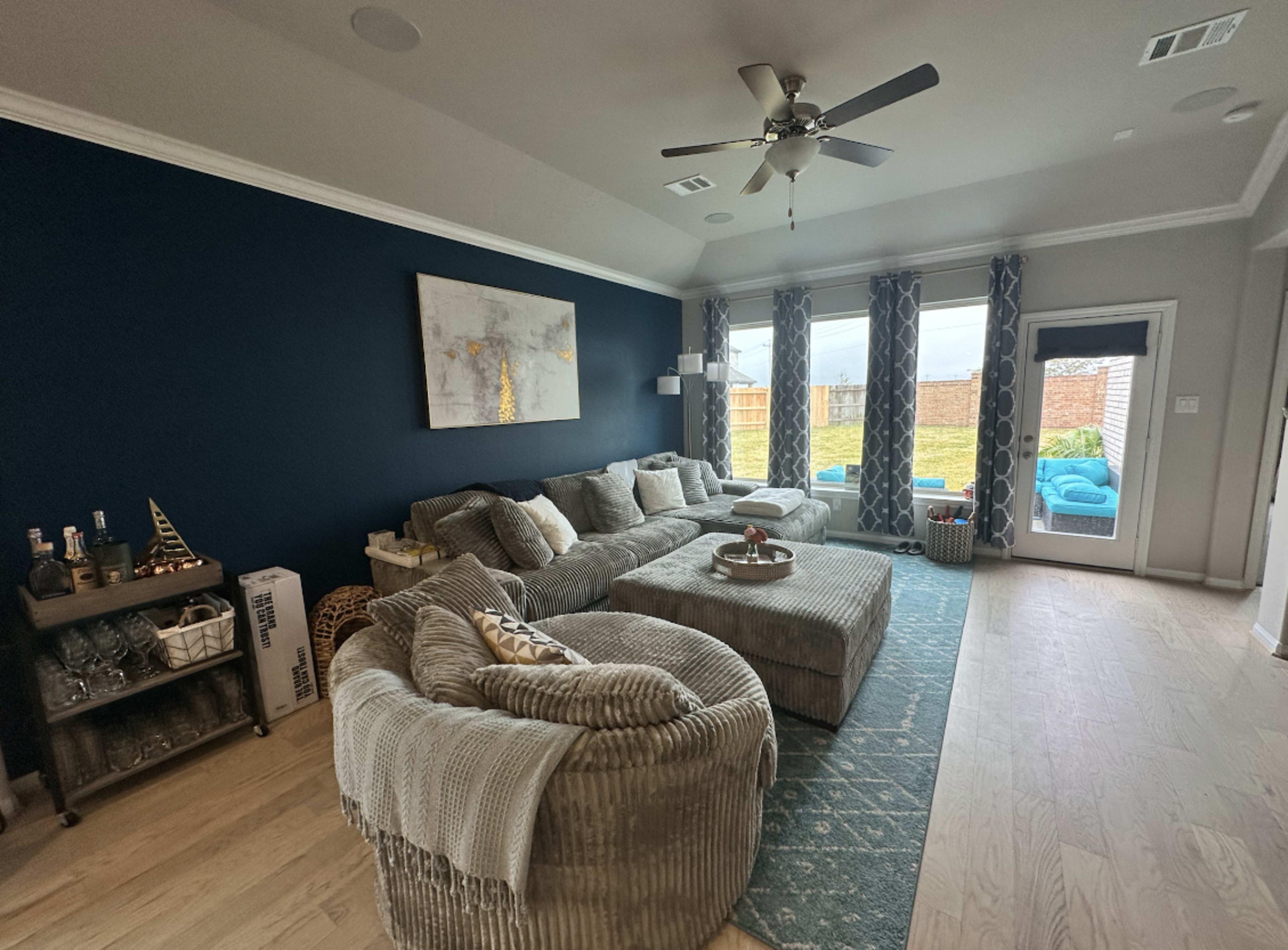 A modern living room features a large sectional sofa, a coffee table, and a stylish area rug, with large windows overlooking a backyard.