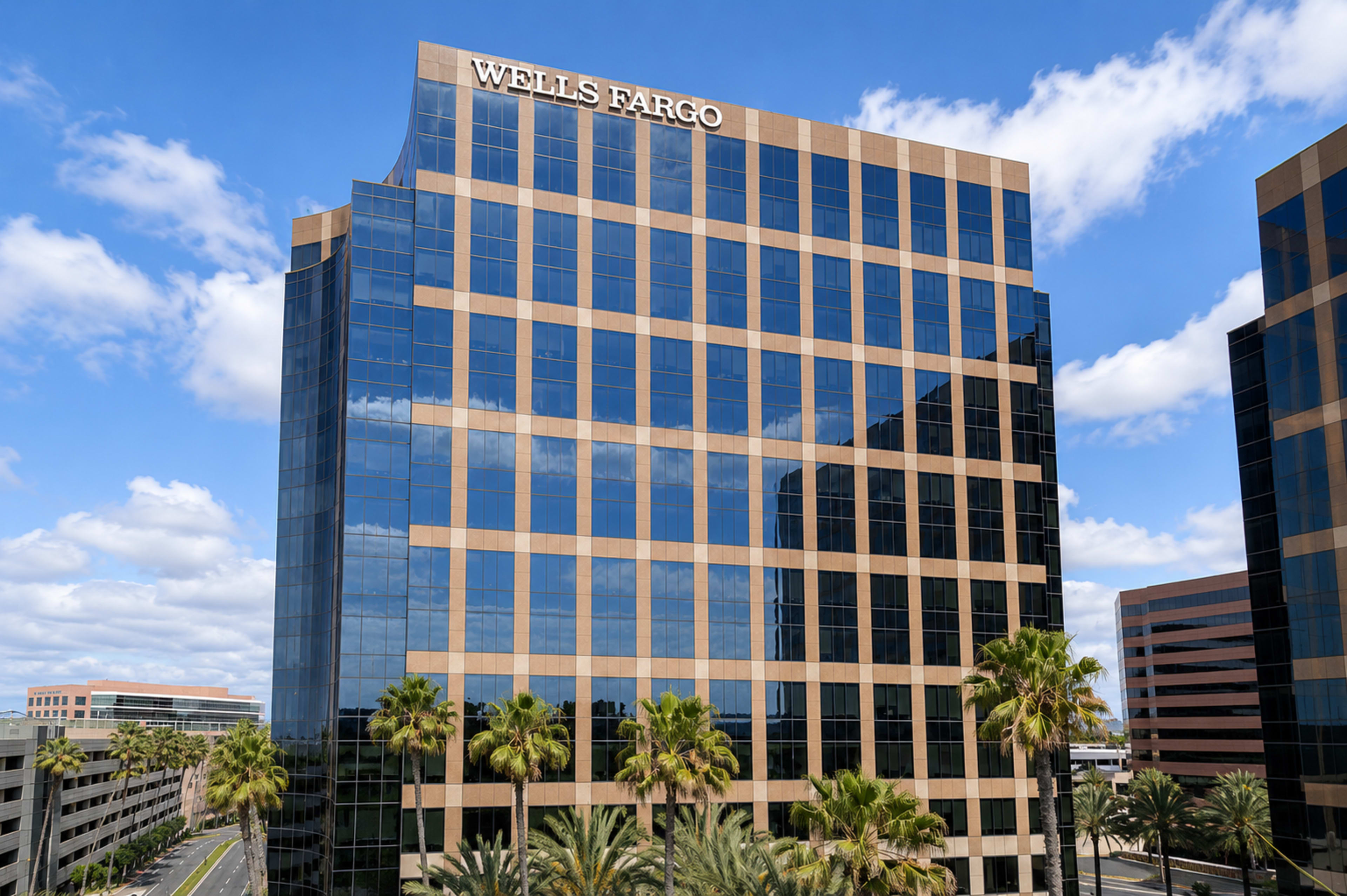 The image shows a tall Wells Fargo office building with a glass facade surrounded by palm trees under a partly cloudy sky.
