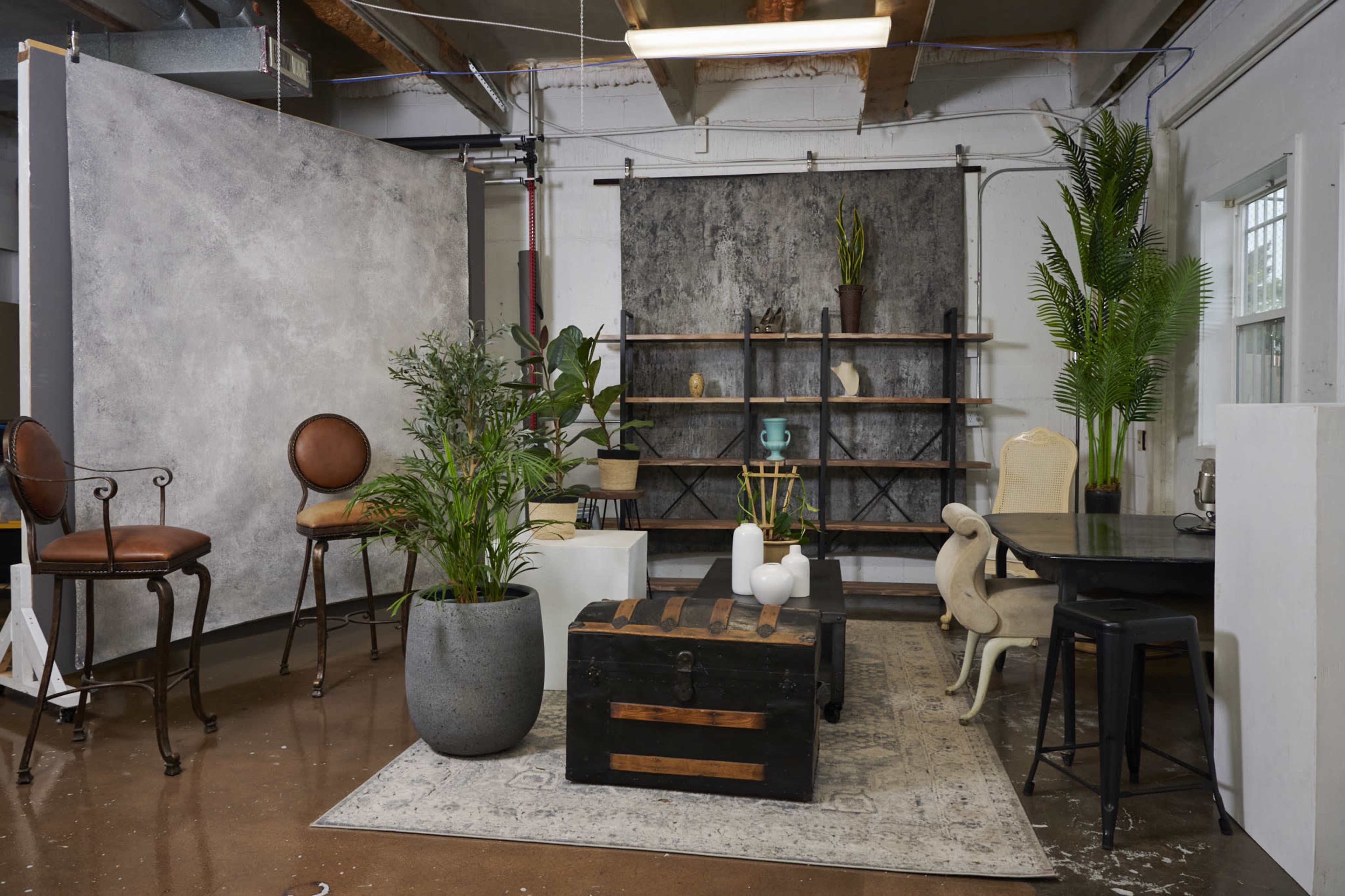 The image shows an interior studio space decorated with a mix of furniture, plants, and backdrops, including a wooden table, chairs, shelves, and various decorative elements.