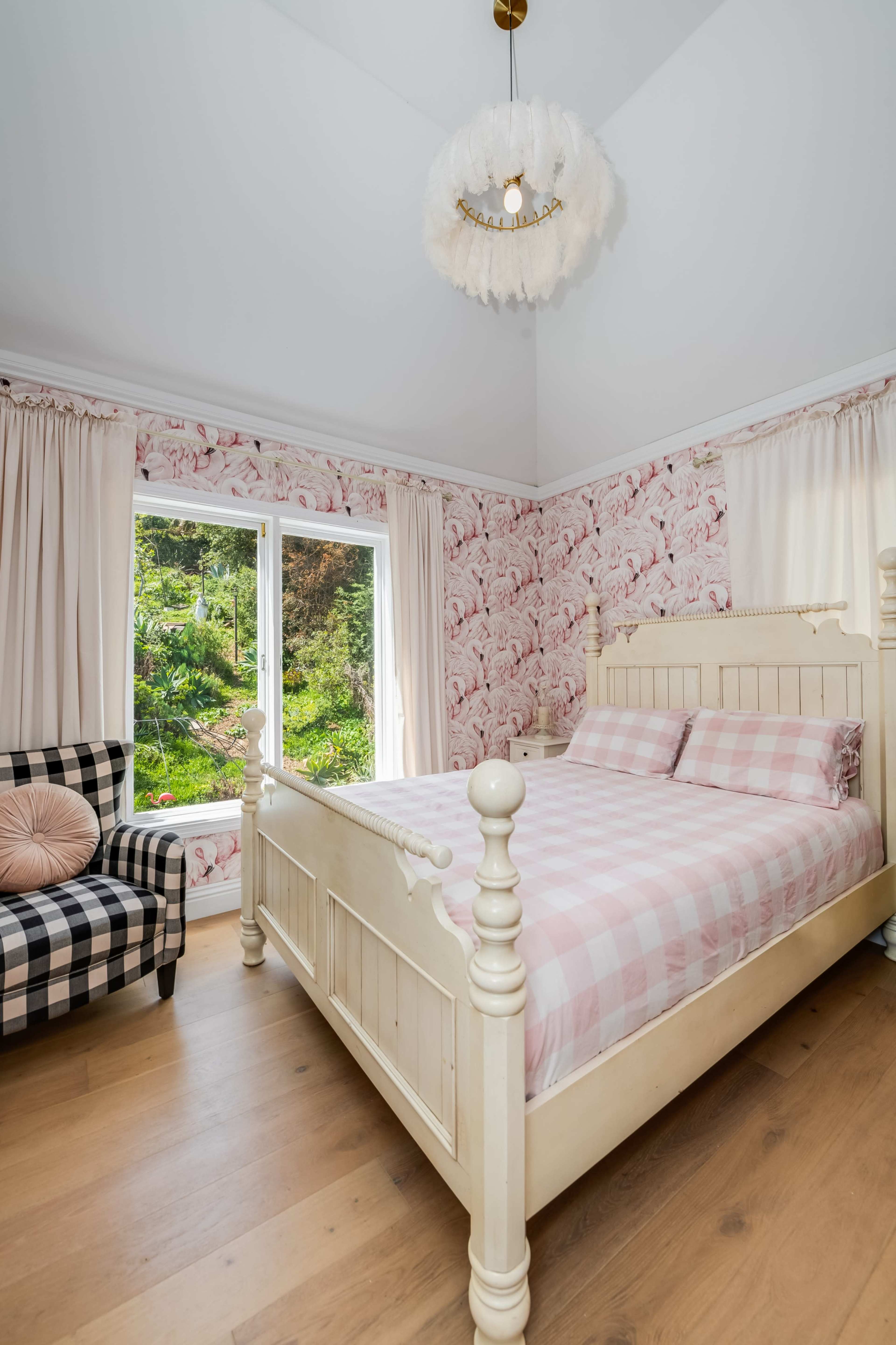 A cozy bedroom features a pink checkered bed, a patterned armchair, and floral wallpaper, with large windows offering a view of greenery outside.