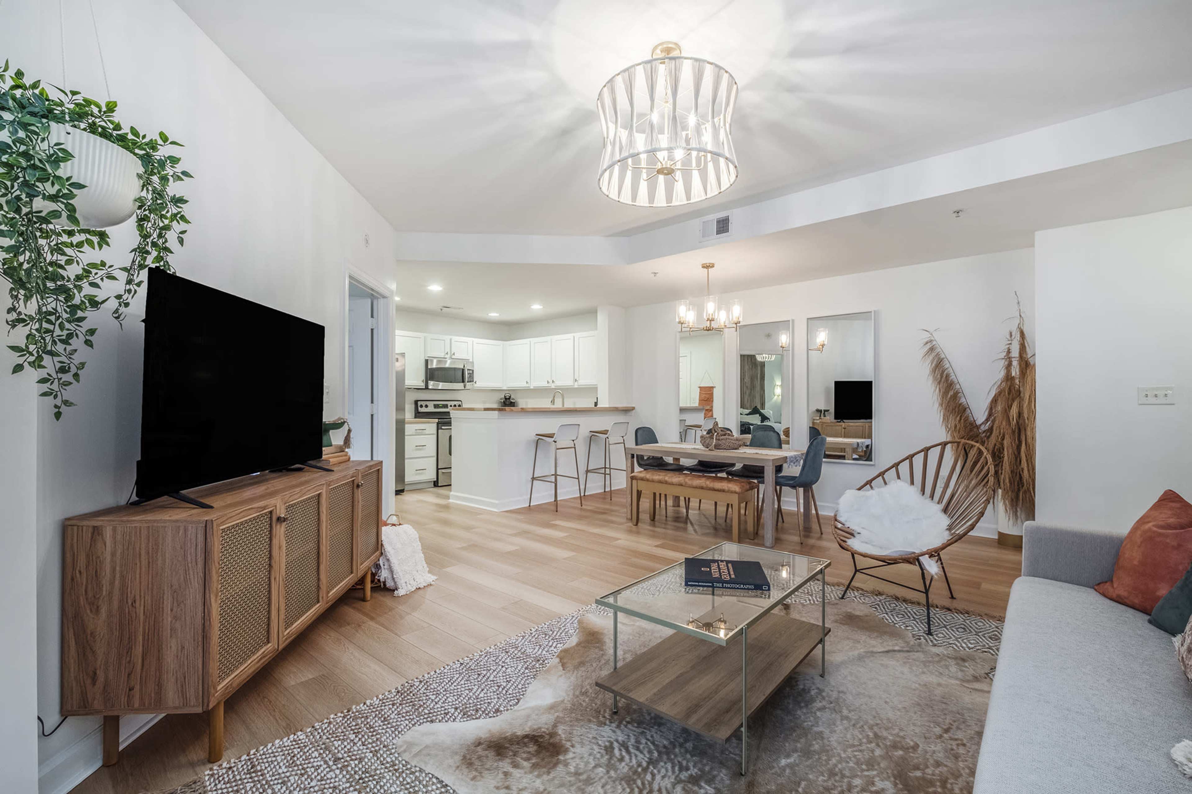 A modern living room features a television stand, a glass coffee table, a sectional sofa, and a dining area with a kitchen in the background.