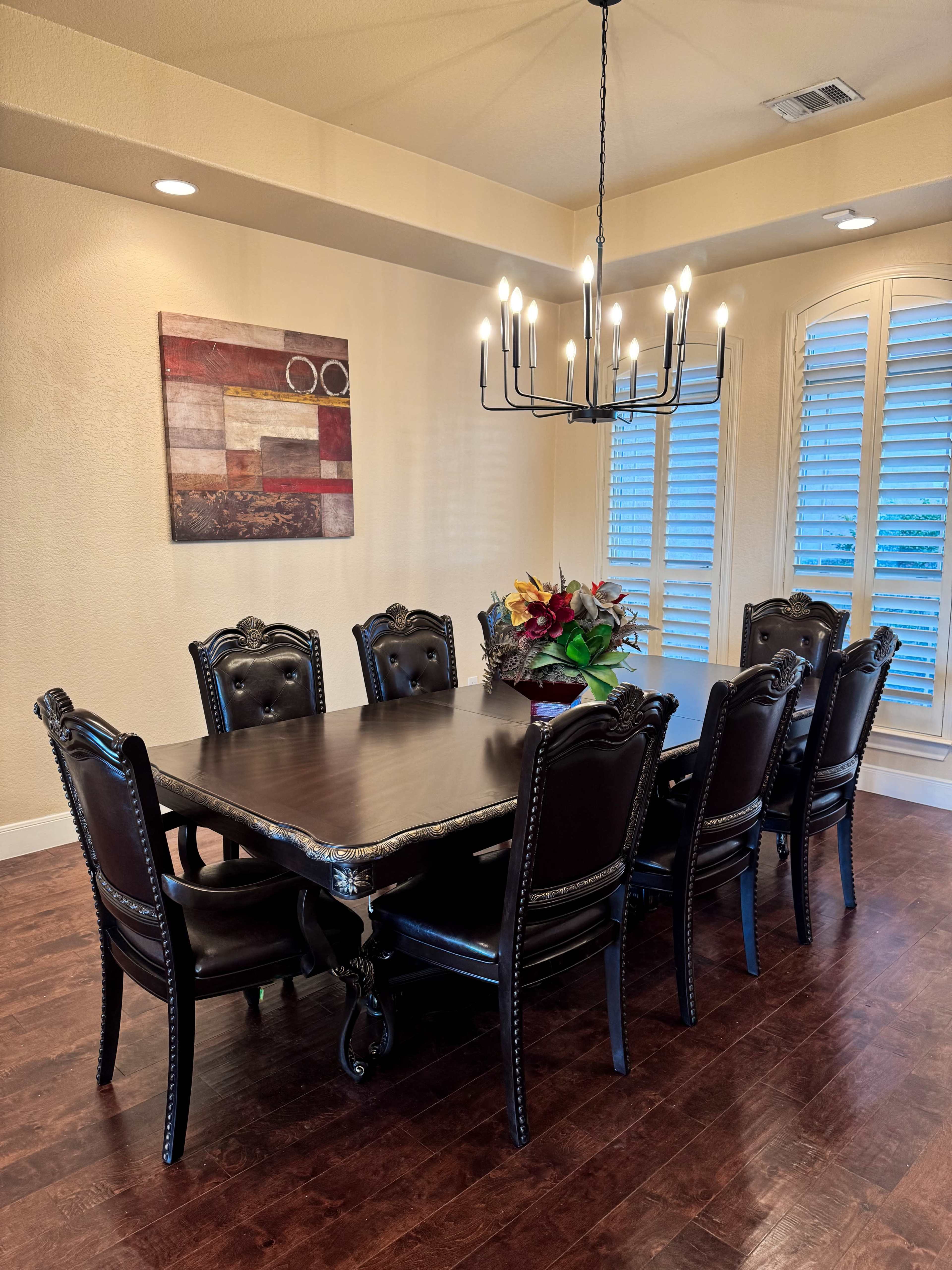 A large, dark wooden dining table with eight upholstered chairs is set in a brightly lit dining room, adorned with a chandelier and a piece of wall art.