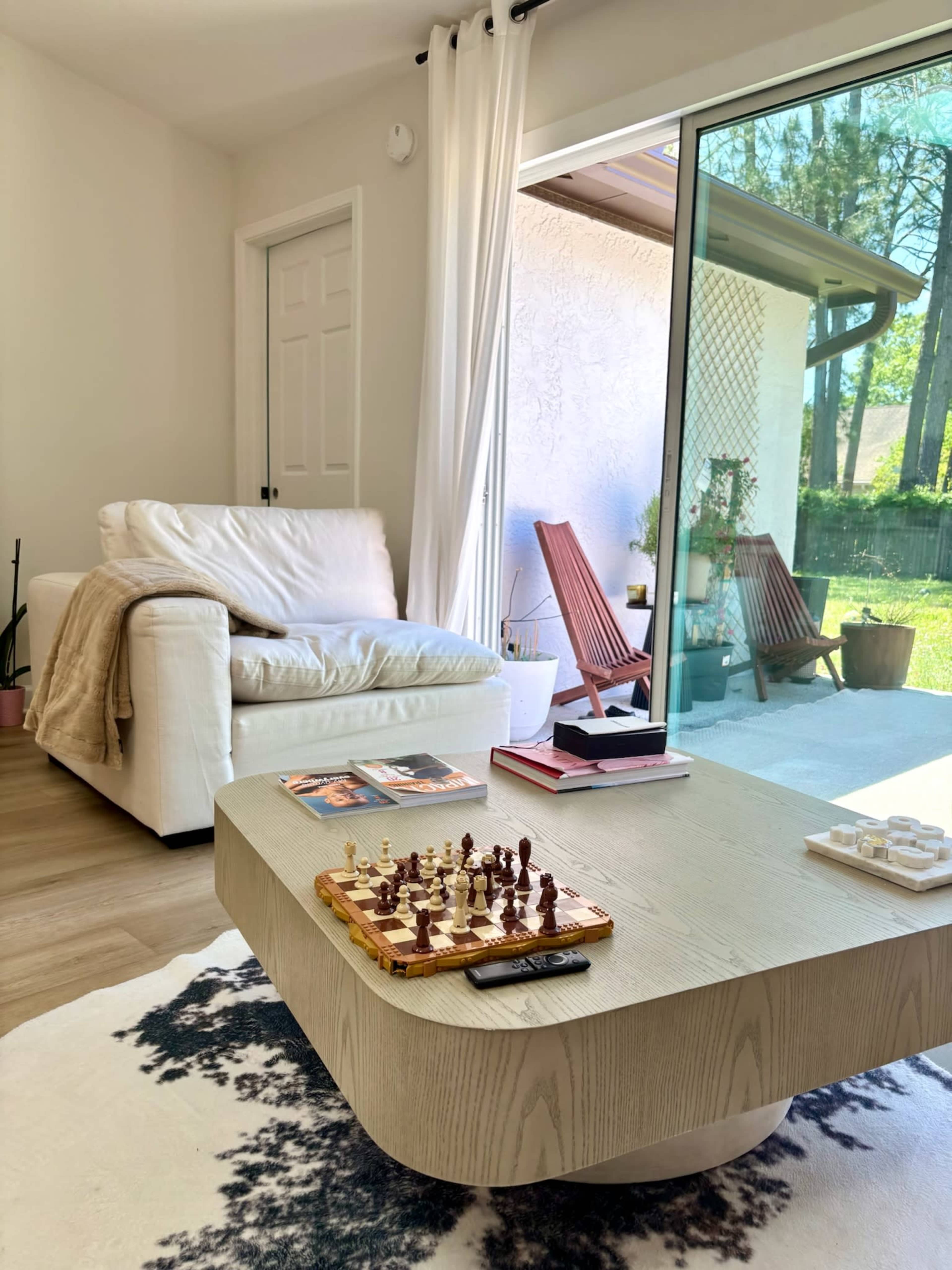 The image shows a modern living room featuring a light-colored sofa, a low coffee table with a chess set, and a large glass door leading to an outdoor area with two wooden chairs.