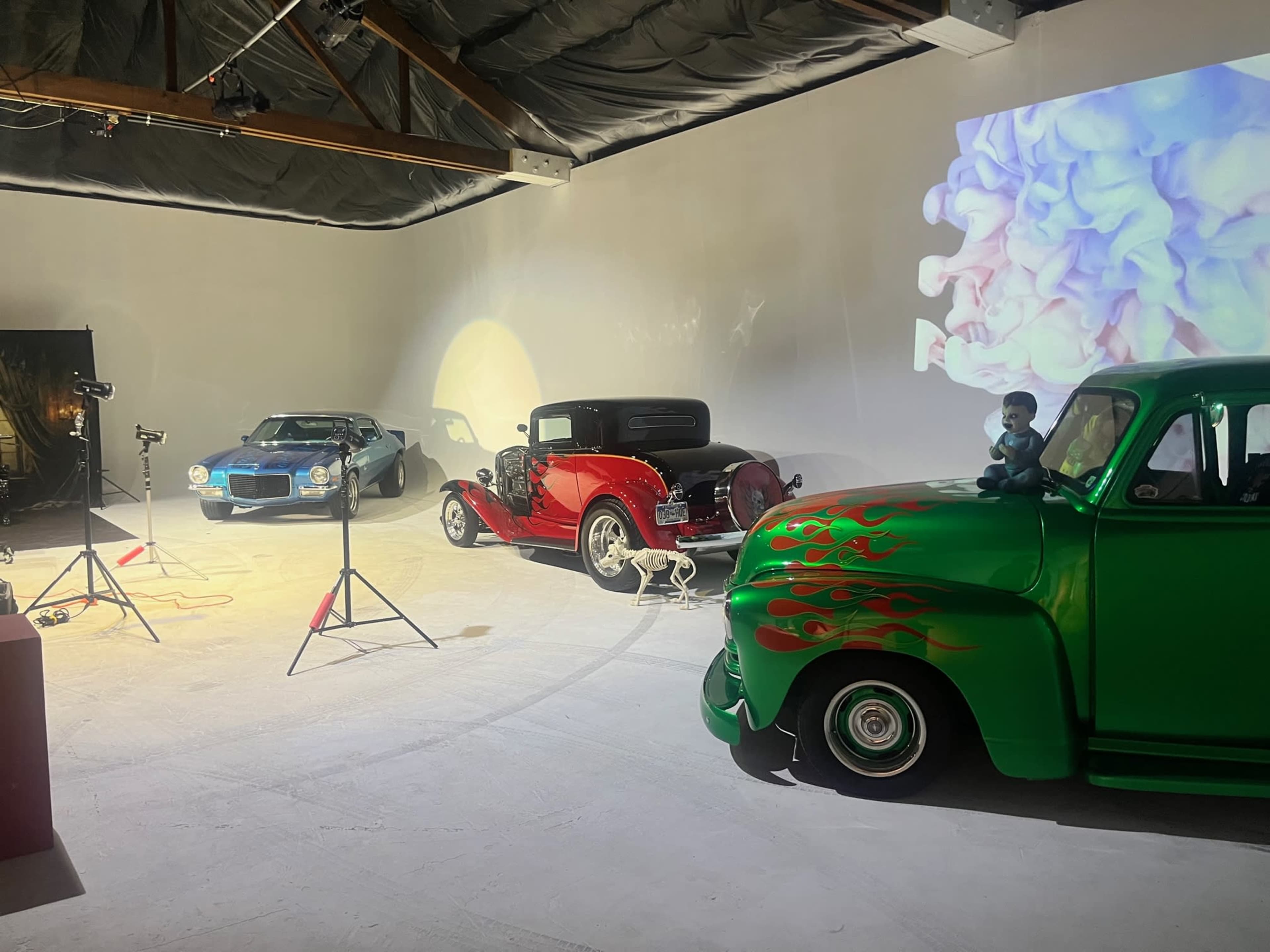The image shows a studio scene with three classic cars—a turquoise car, a red vintage car, and a green truck with flame graphics—alongside photography equipment and a colorful projection on the wall.