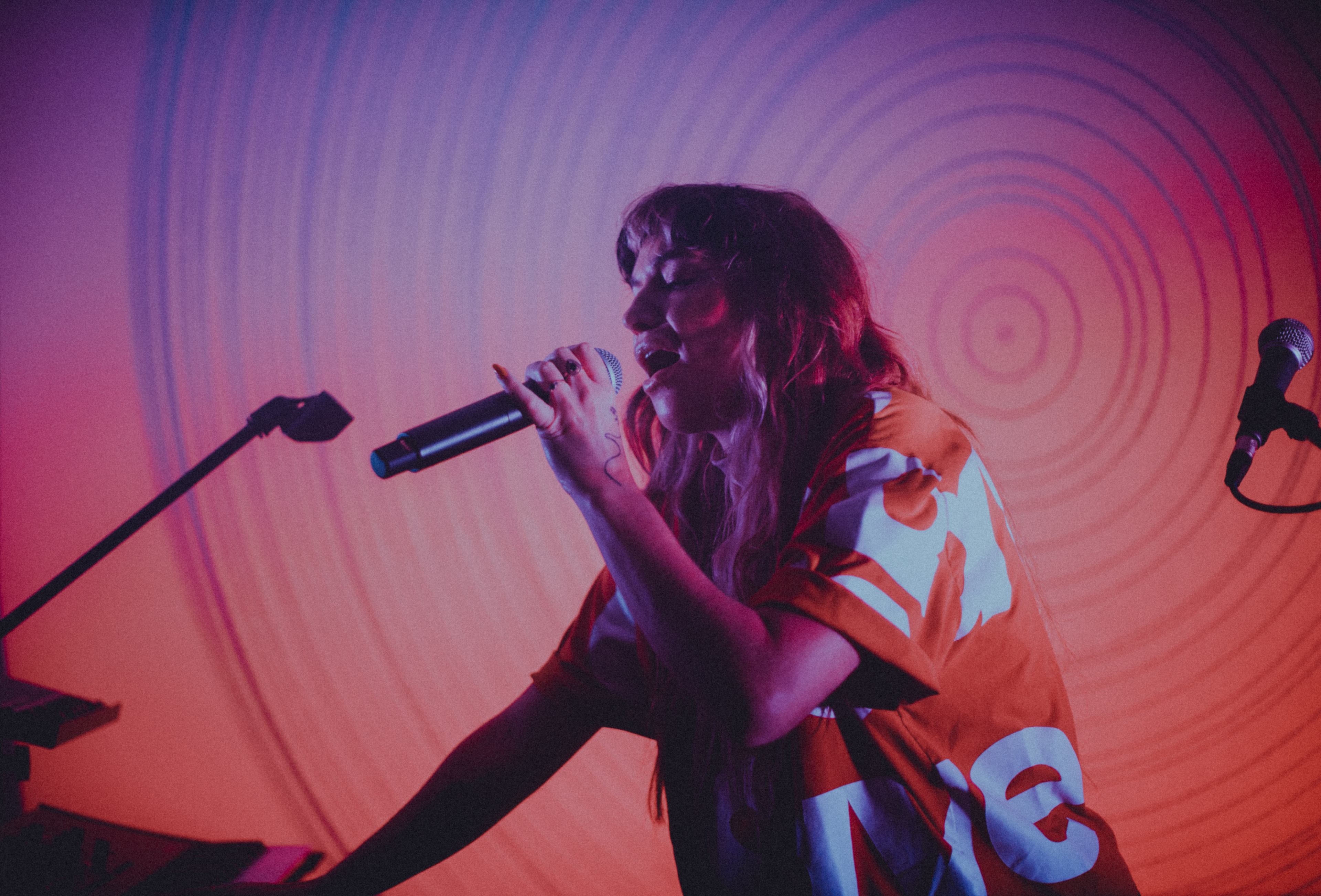 A performer sings into a microphone on stage while wearing an oversized shirt, against a backdrop of swirling colors.