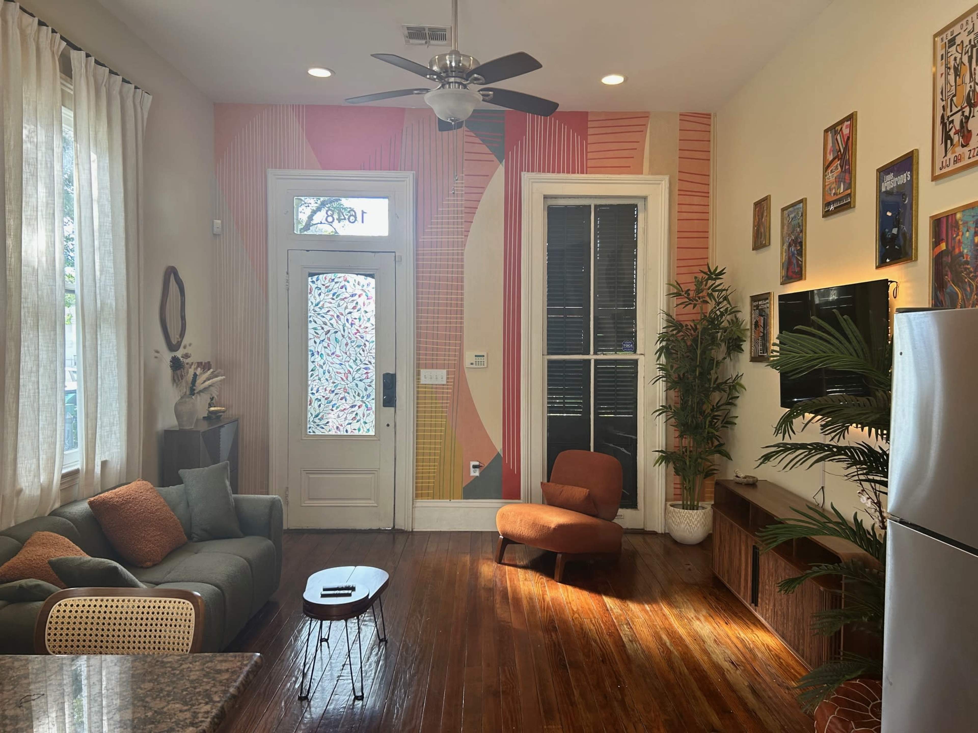 The image shows a cozy living space with a small kitchen area, featuring a colorful accent wall, a door with patterned glass, and a mix of modern furniture.