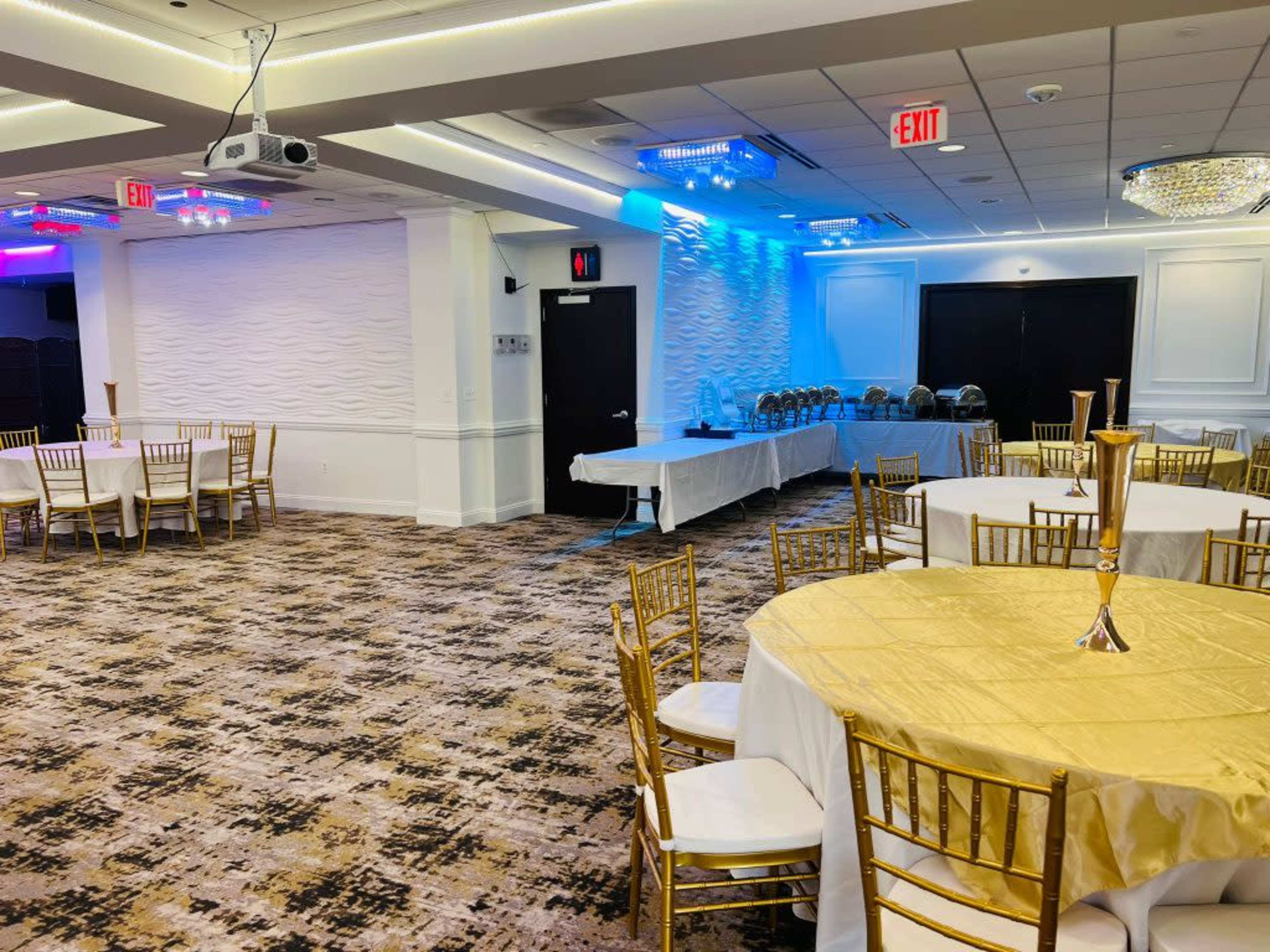 The image shows a banquet hall set up with round tables covered in gold tablecloths and a buffet setup against one wall, illuminated by colorful lighting.