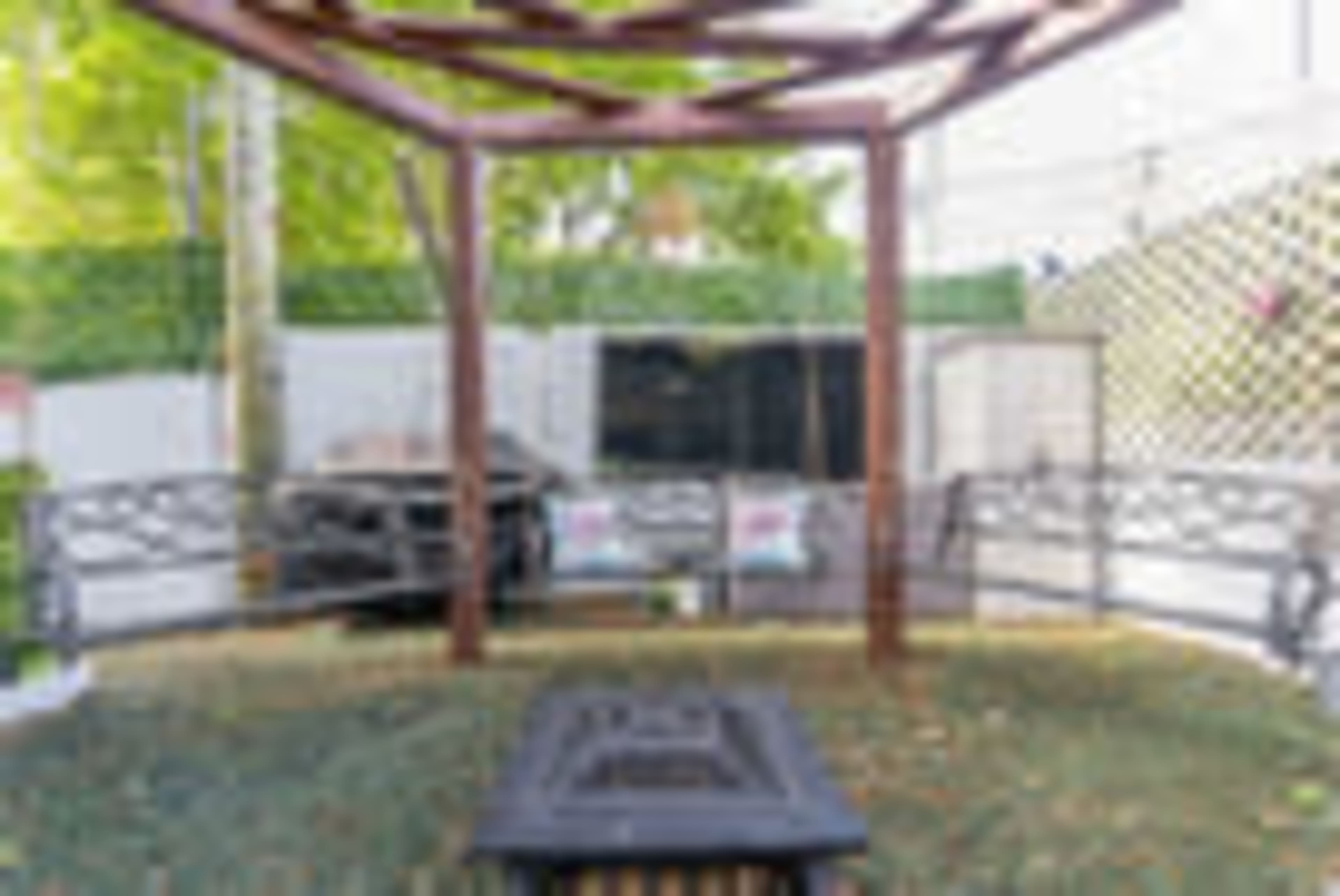 A wooden gazebo with a bench surrounds a circular fire pit, set in a landscaped outdoor area.