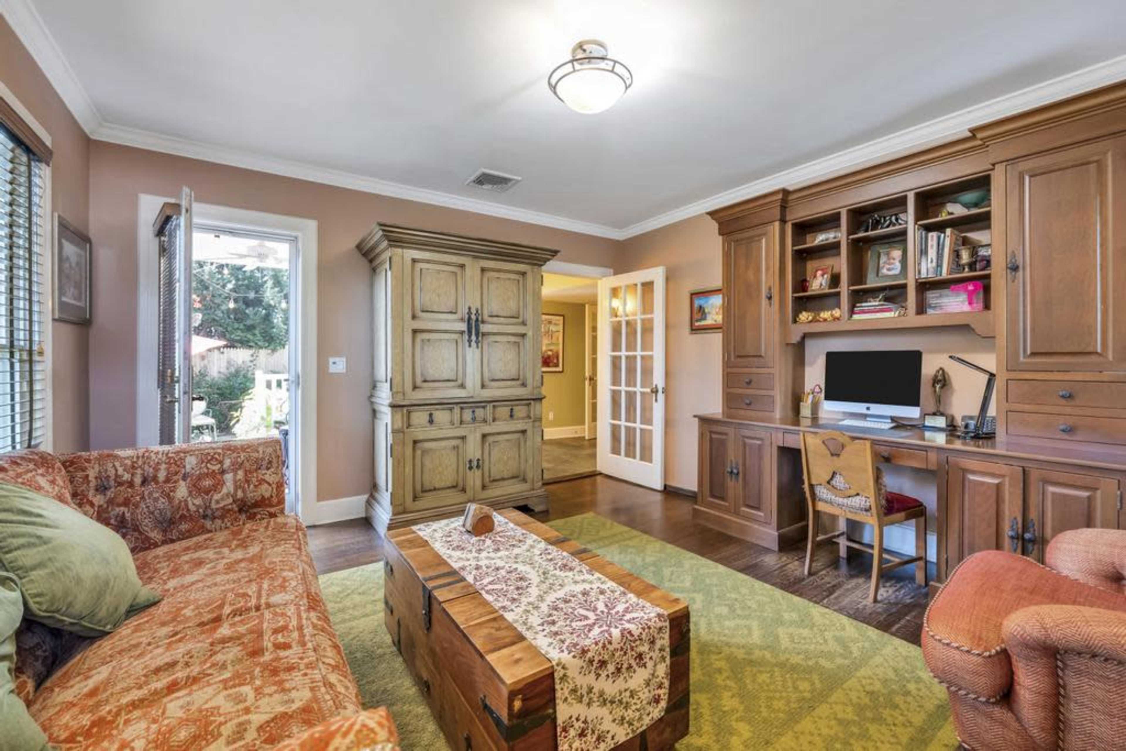 A cozy living room featuring a patterned sofa, a wooden coffee table, a desk with a computer, and a large cabinet, all arranged with natural light filtering in from a nearby doorway.