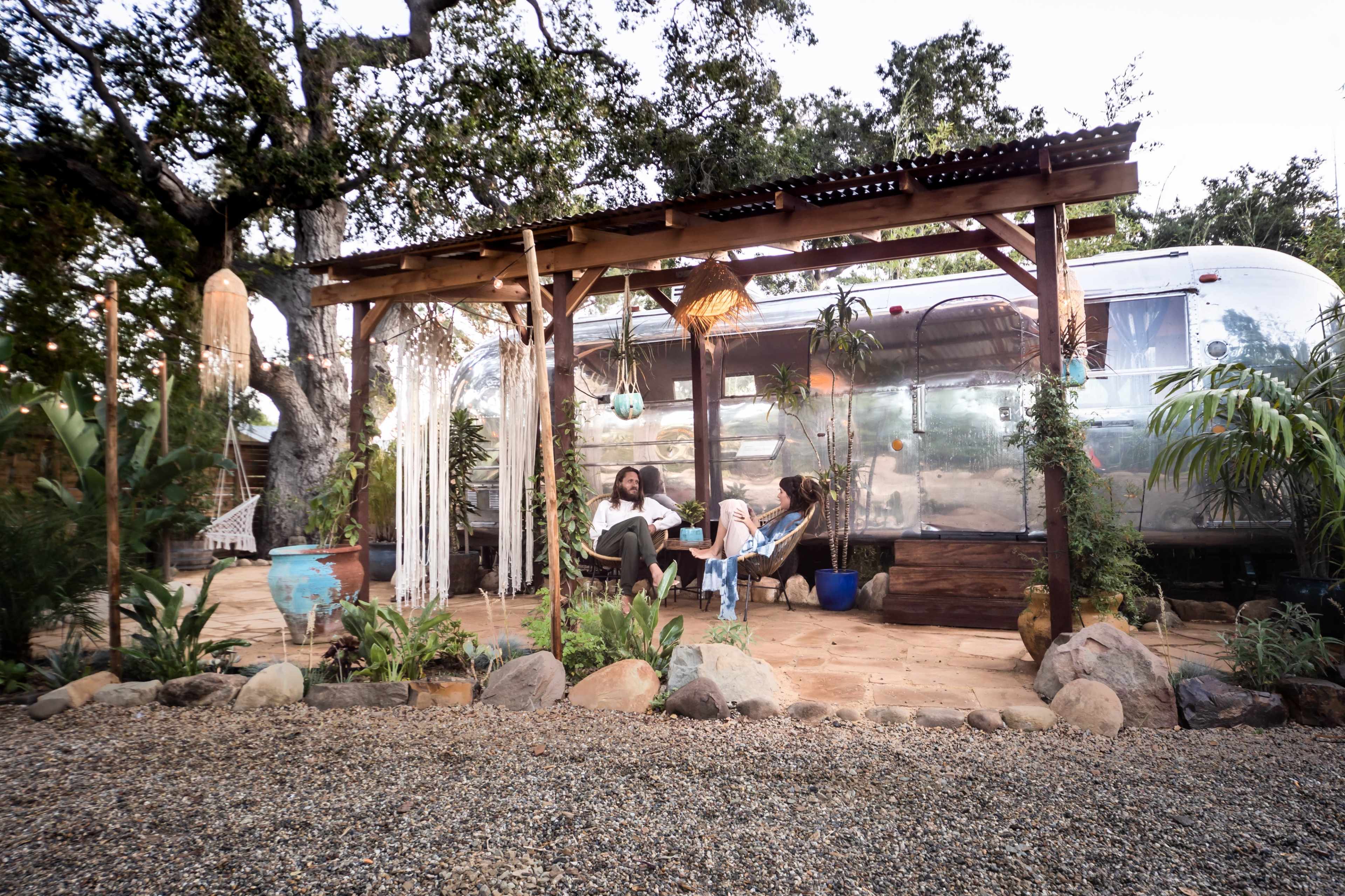 Two people relax under a wooden pergola adorned with plants, in front of a silver Airstream trailer surrounded by gravel and greenery.