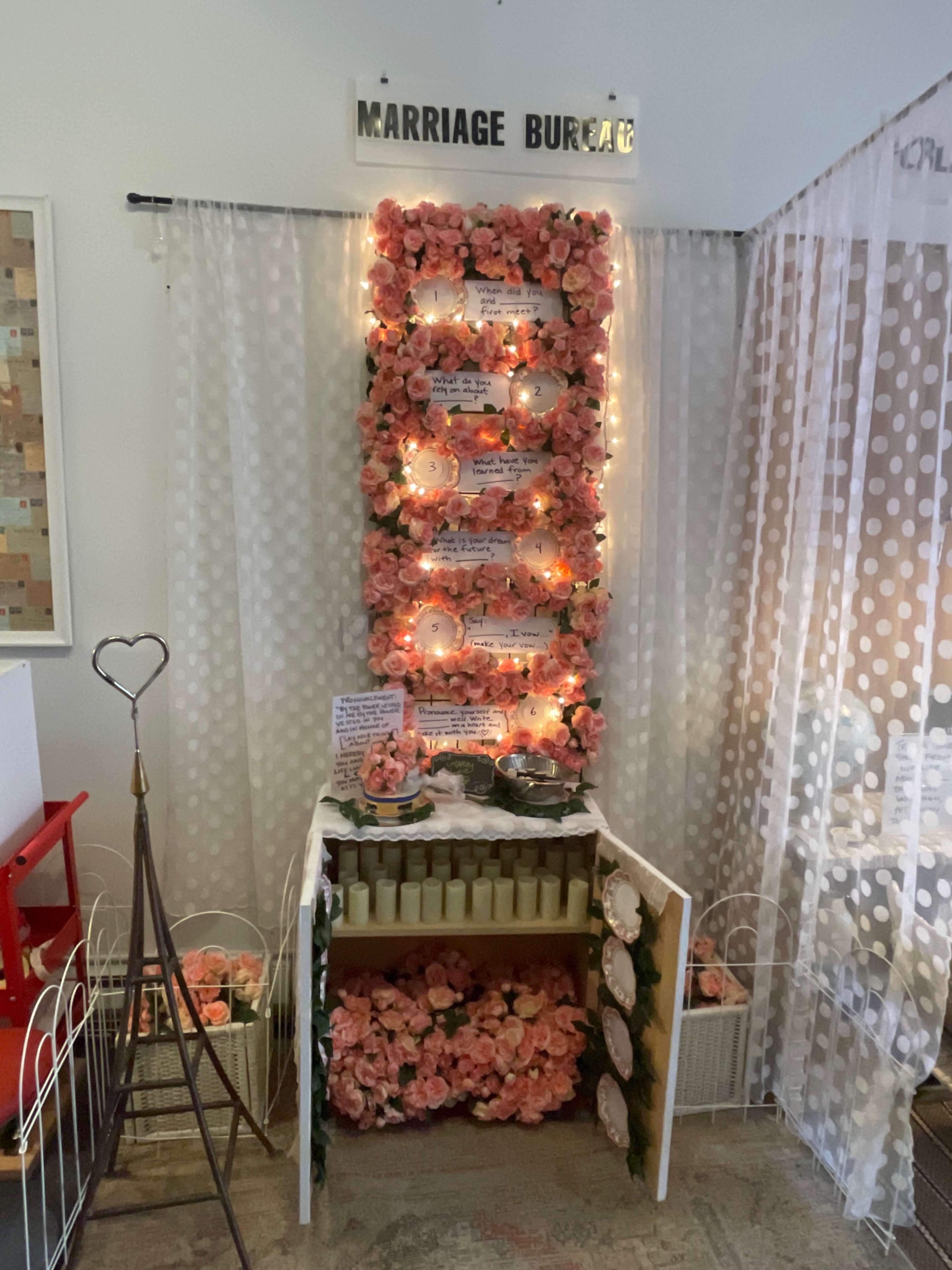 The image shows a decorative display labeled "Marriage Bureau," featuring pink flowers, soft lighting, and several signs or messages arranged on a vertical board.