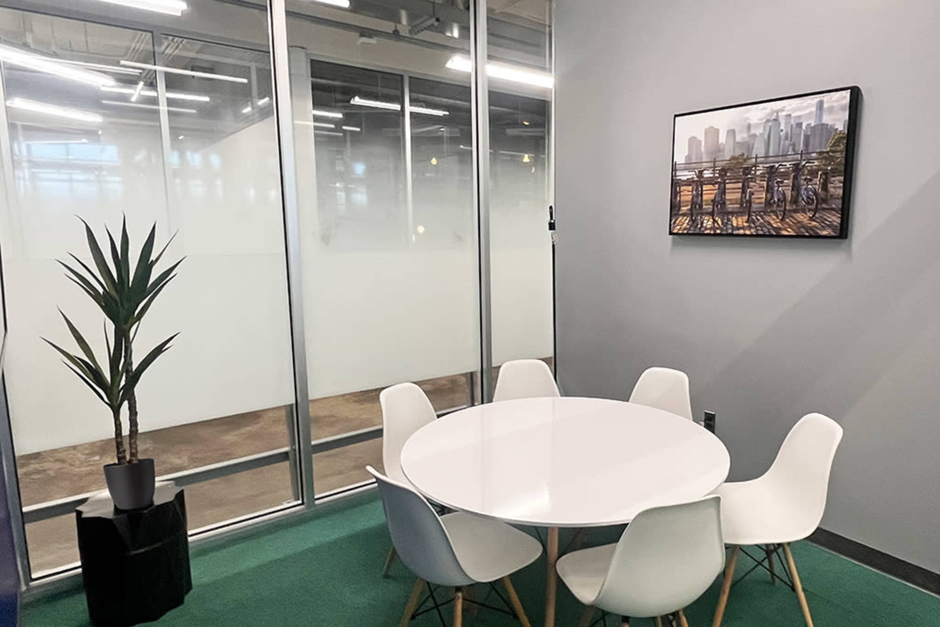 A round white table surrounded by six chairs is set in a glass-walled conference room featuring a plant and a cityscape painting on the wall.