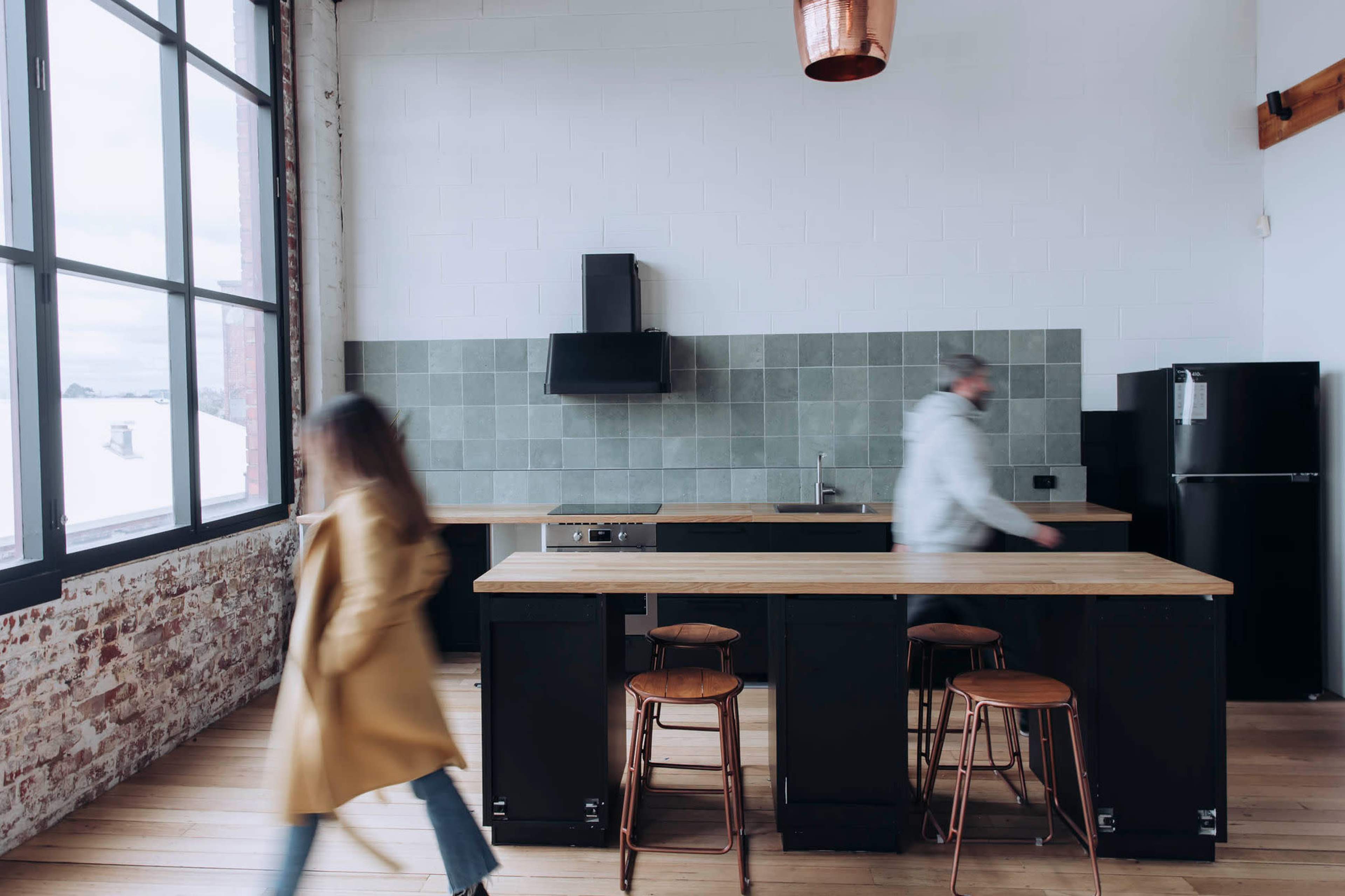 A kitchen features a large countertop with three wooden stools, a black refrigerator, and a person walking past while another stands at the counter.