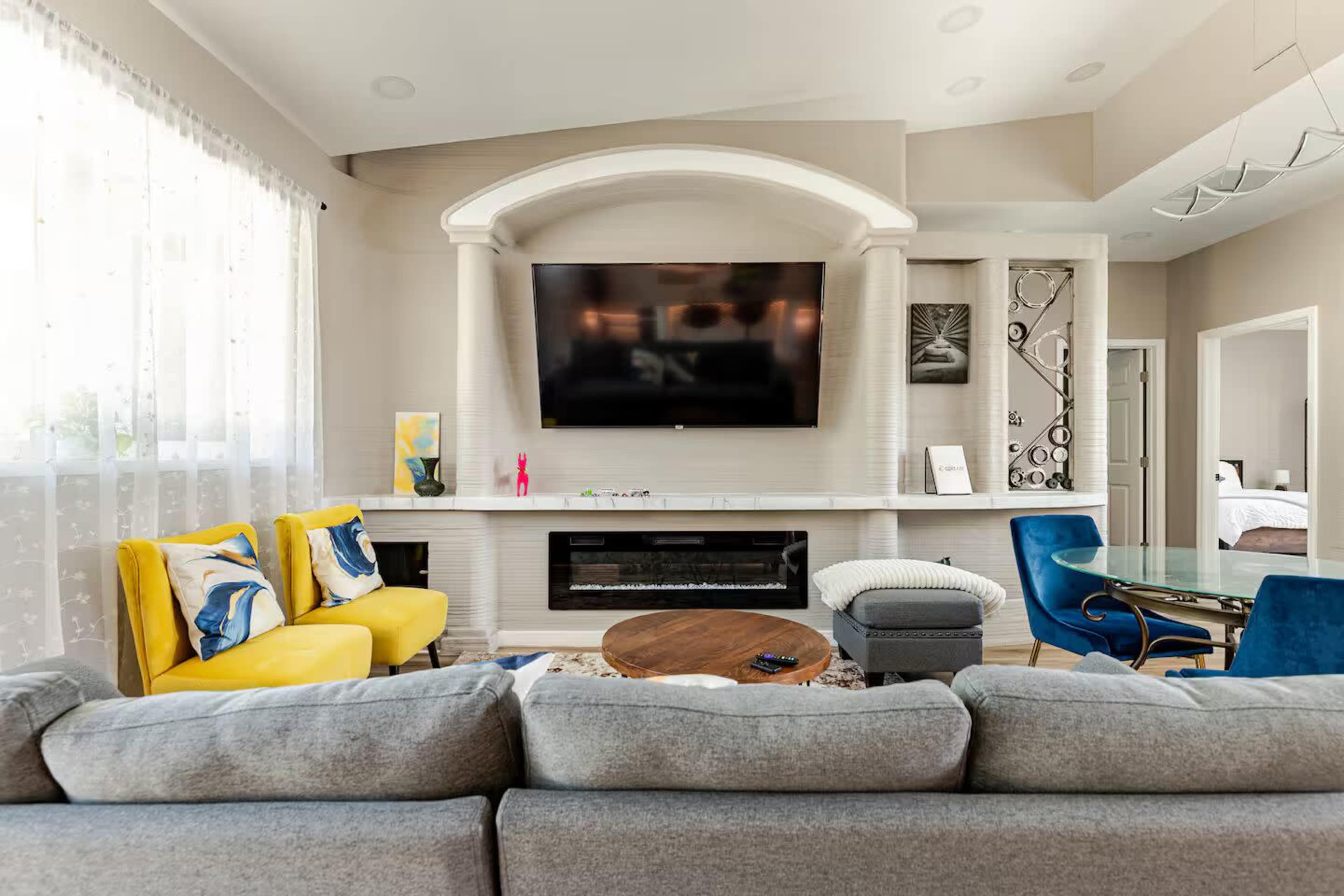 The living room features a large flat-screen TV mounted above a modern fireplace, with a gray sofa, yellow accent chairs, and a glass dining table.