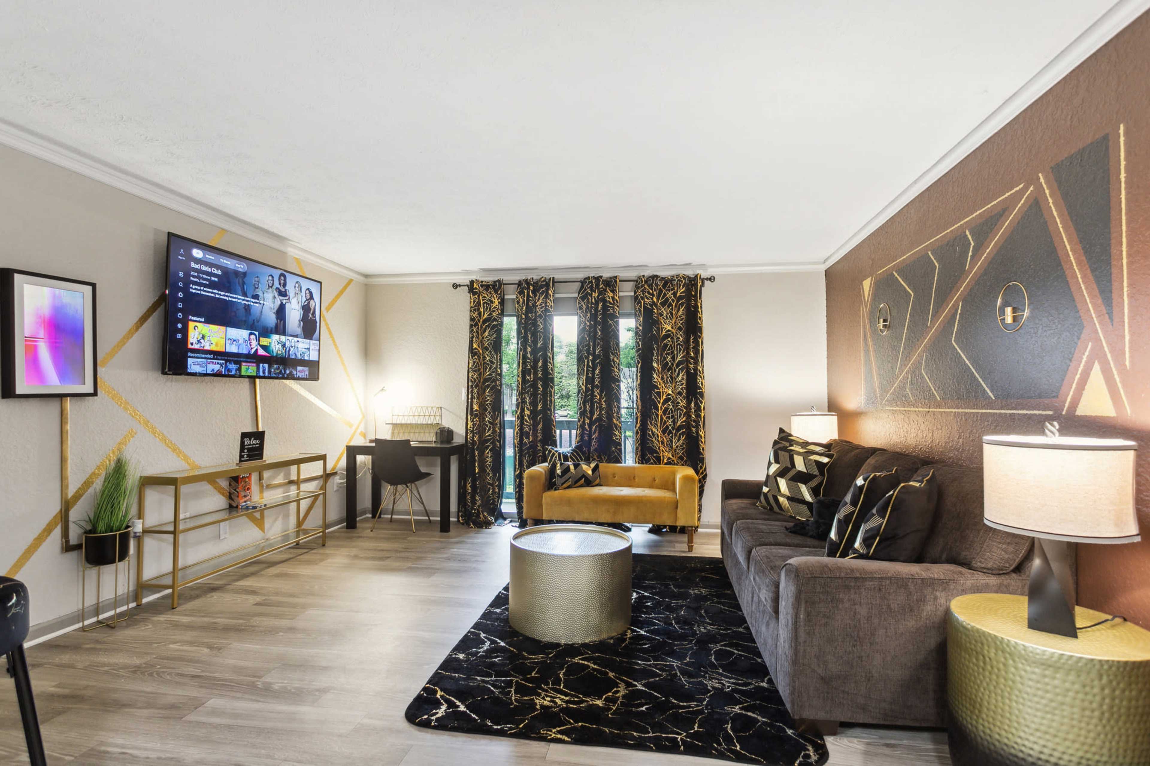 The living room features a modern design with a sectional sofa, gold accents, a circular coffee table, and a television mounted on the wall.