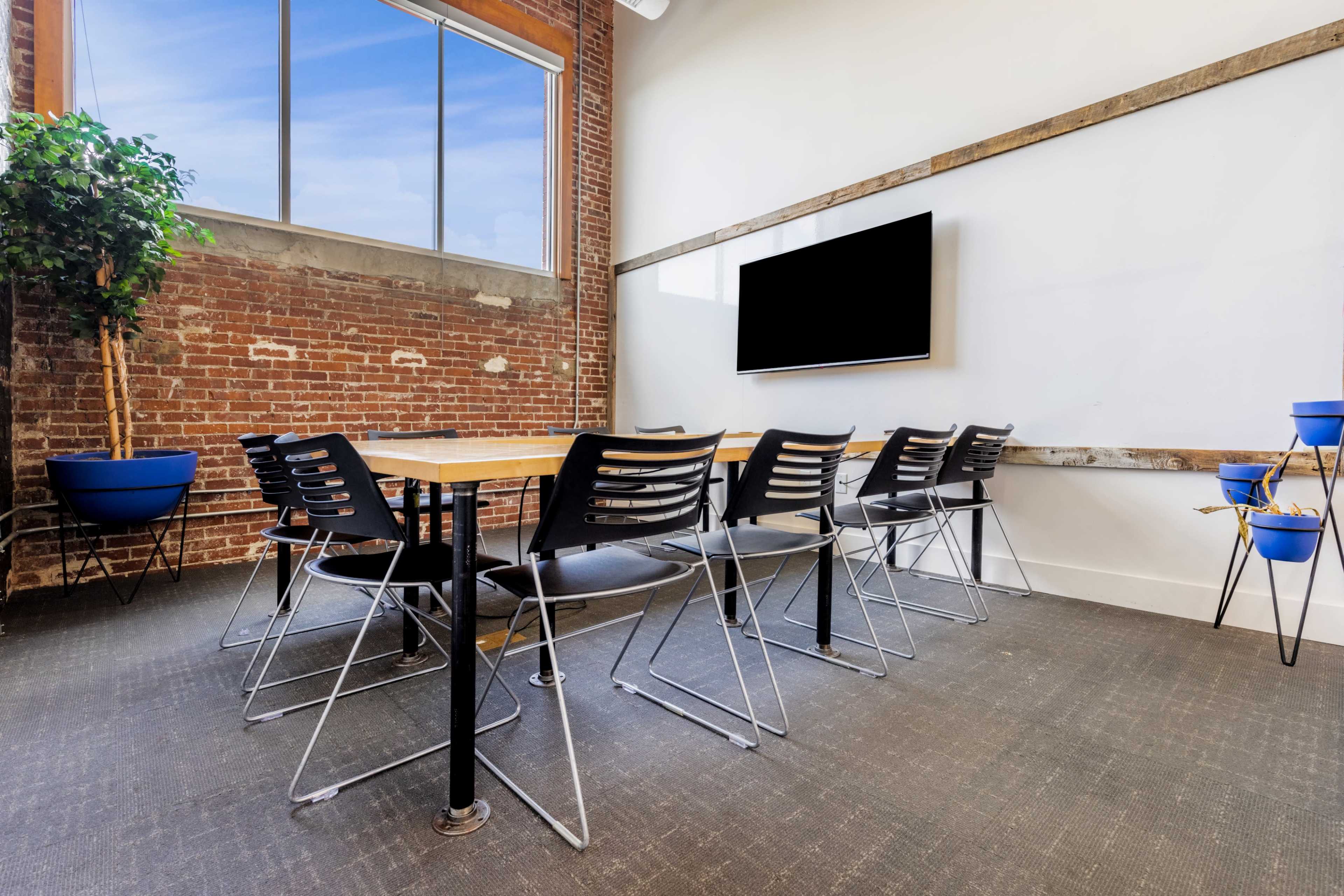 The image shows a conference room with a large wooden table surrounded by black chairs, a television mounted on the wall, and a tall potted plant in the corner.