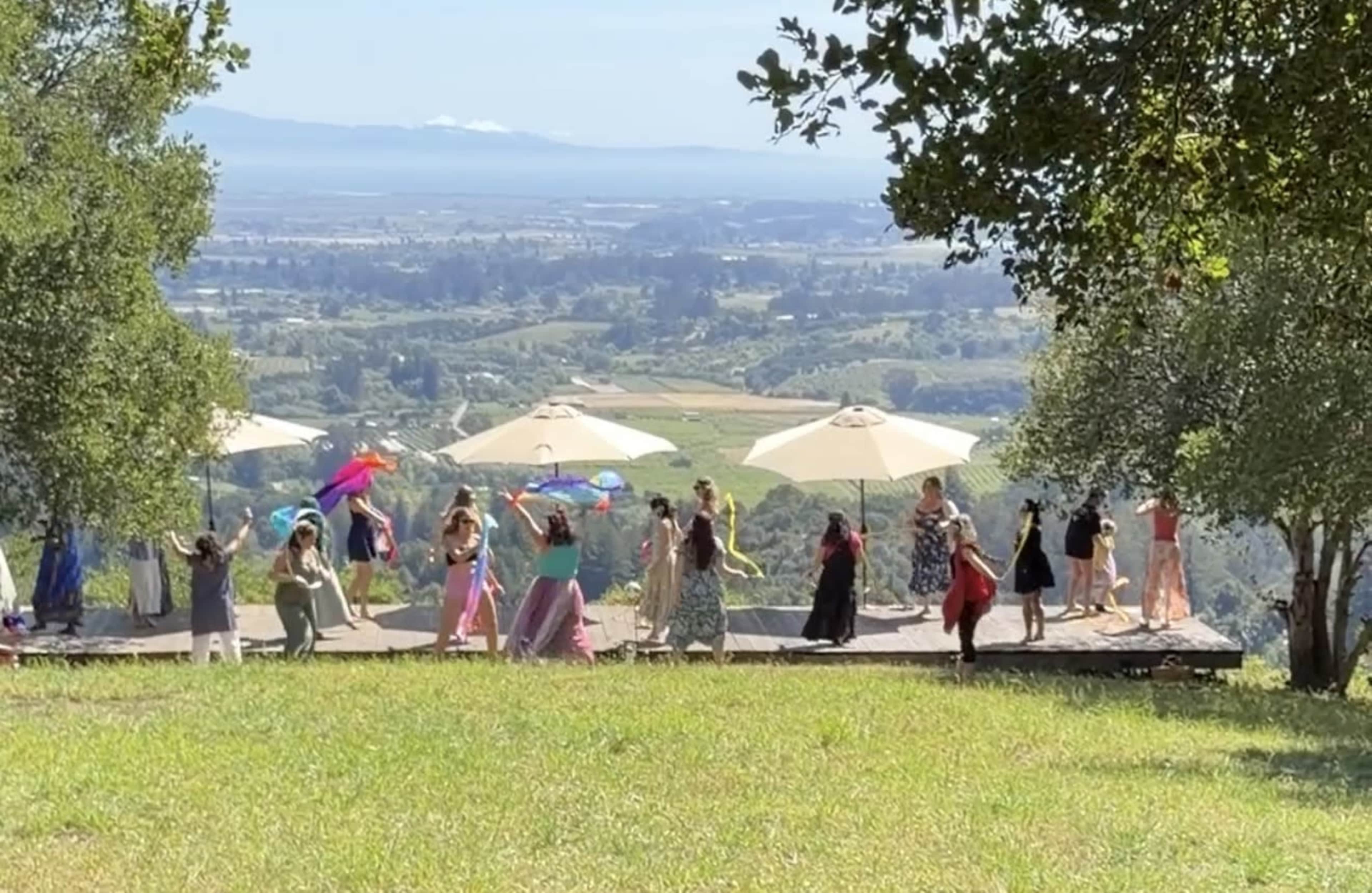 A group of people in flowing dresses dance on a wooden deck surrounded by trees, with umbrellas overhead and a view of a valley in the background.