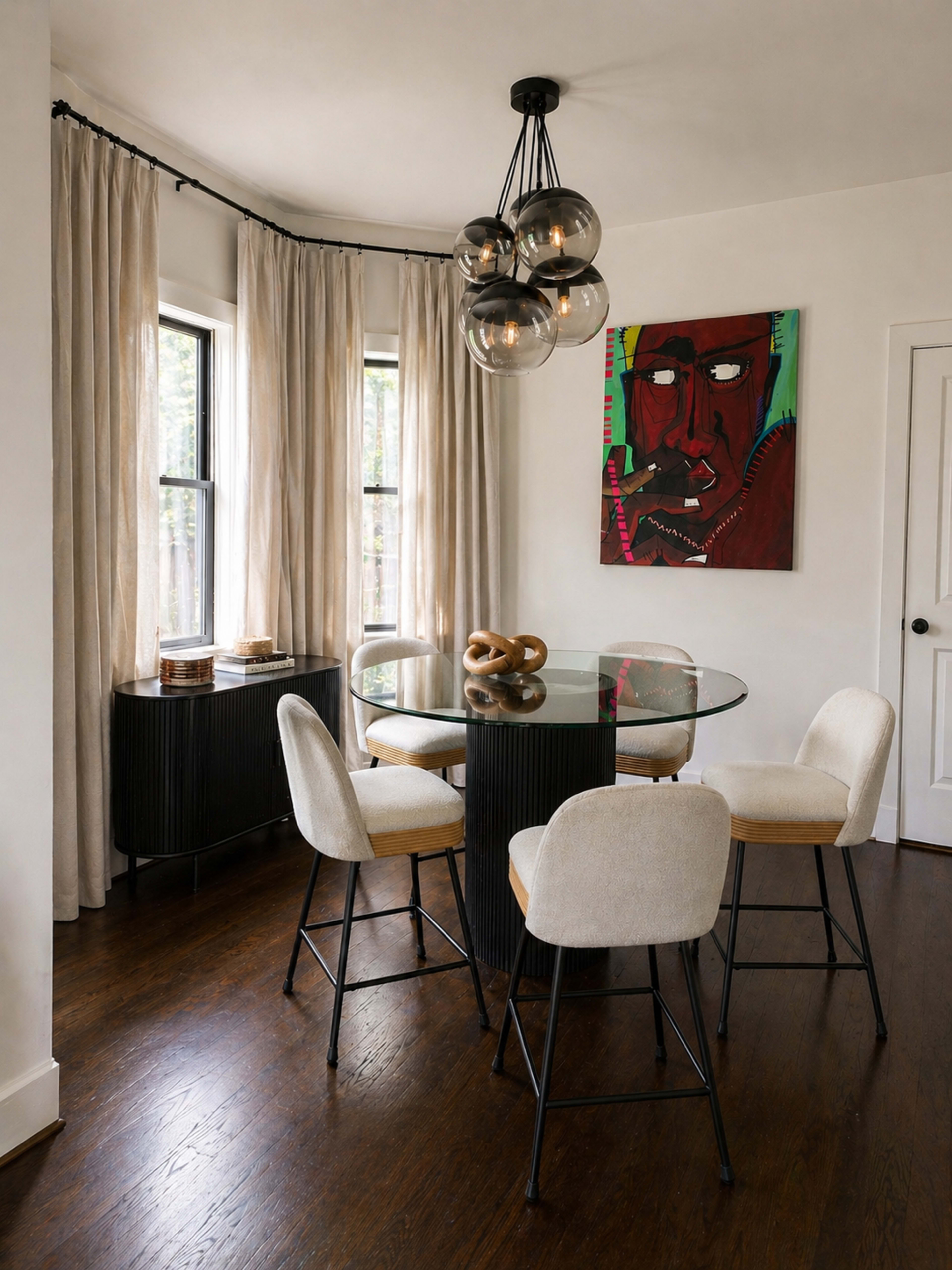 A round glass dining table with four upholstered chairs is positioned in a bright dining area featuring large windows and a colorful artwork on the wall.