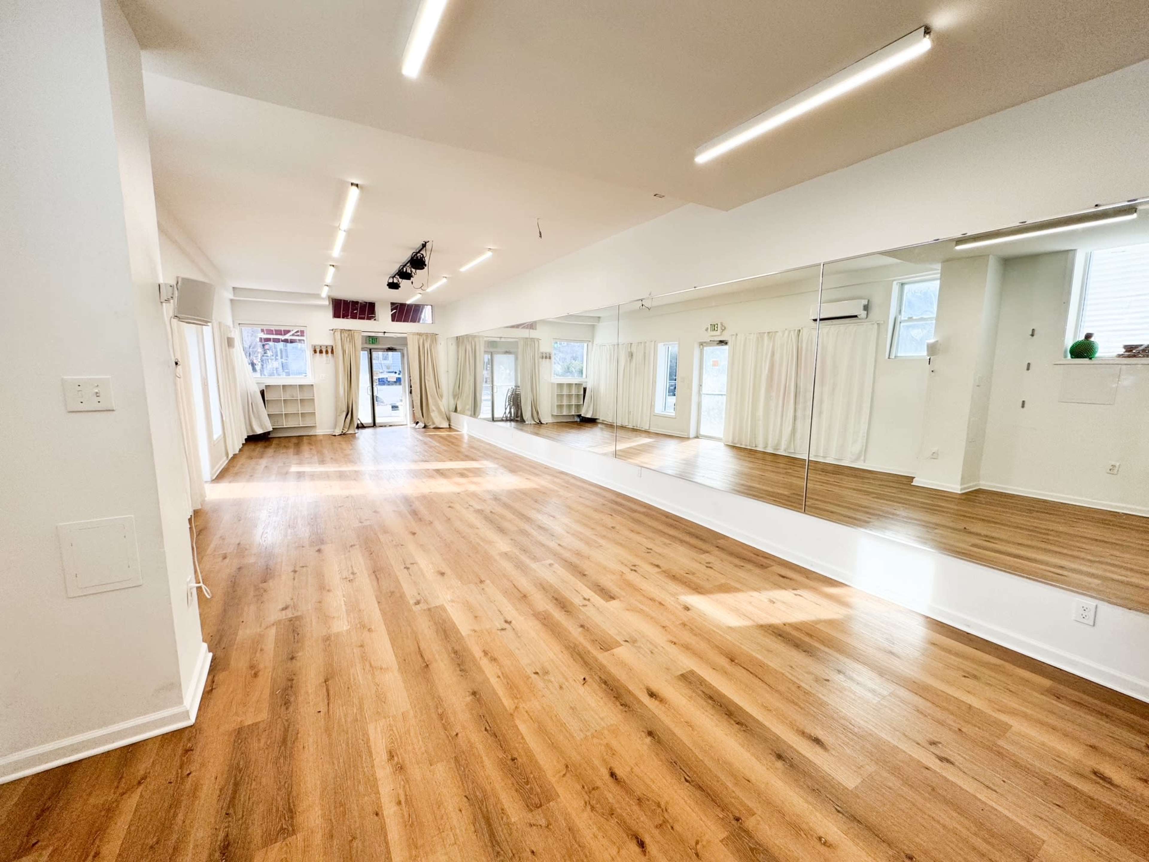 The image depicts a spacious, well-lit studio with wooden flooring, large mirrors on one wall, and natural light streaming through windows.