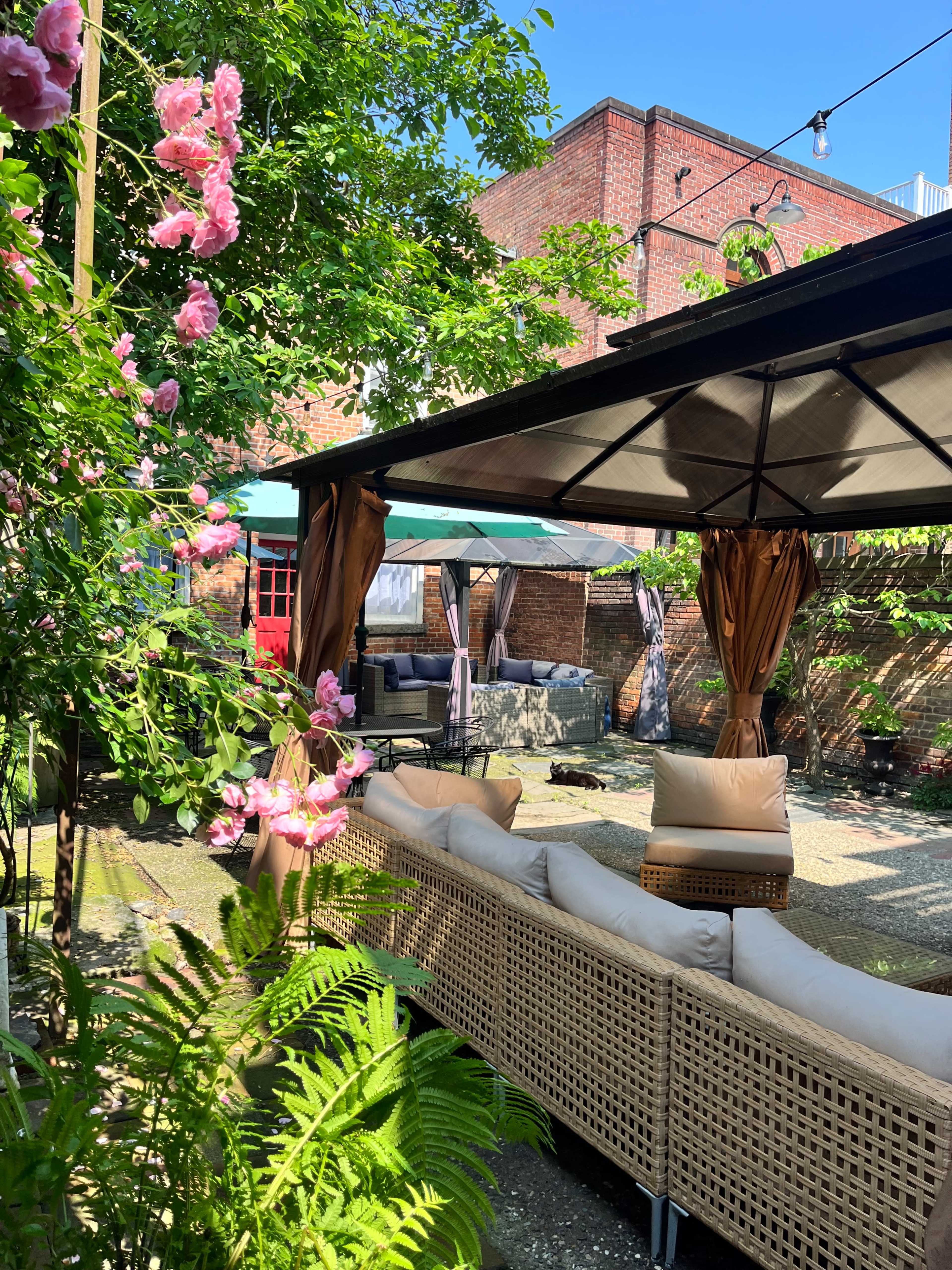 The image shows a cozy outdoor seating area surrounded by greenery and blooming flowers, with a covered pavilion and patio furniture.