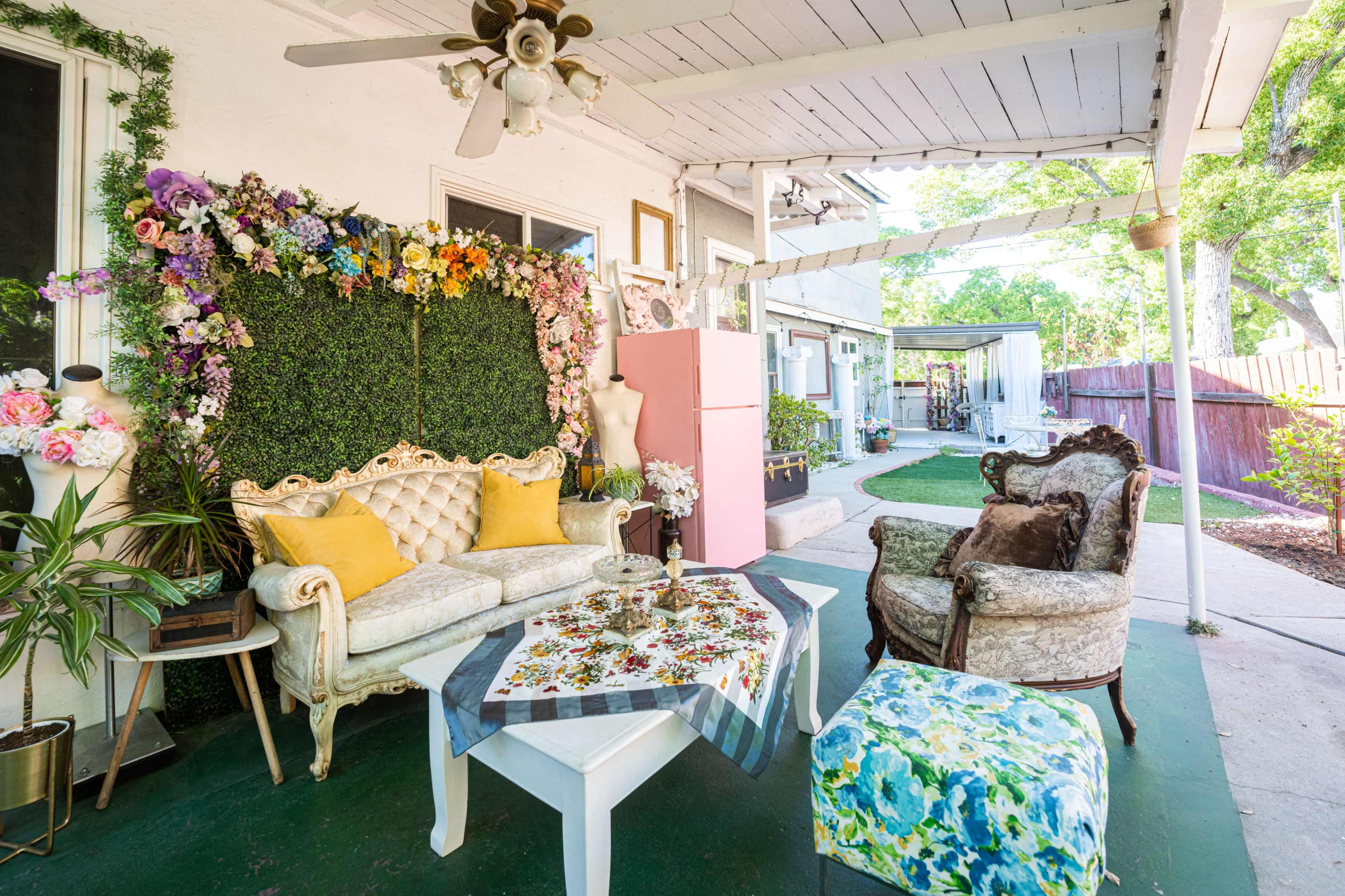 The image shows a patio area with a floral backdrop, vintage furniture, and decorative elements arranged around a table.