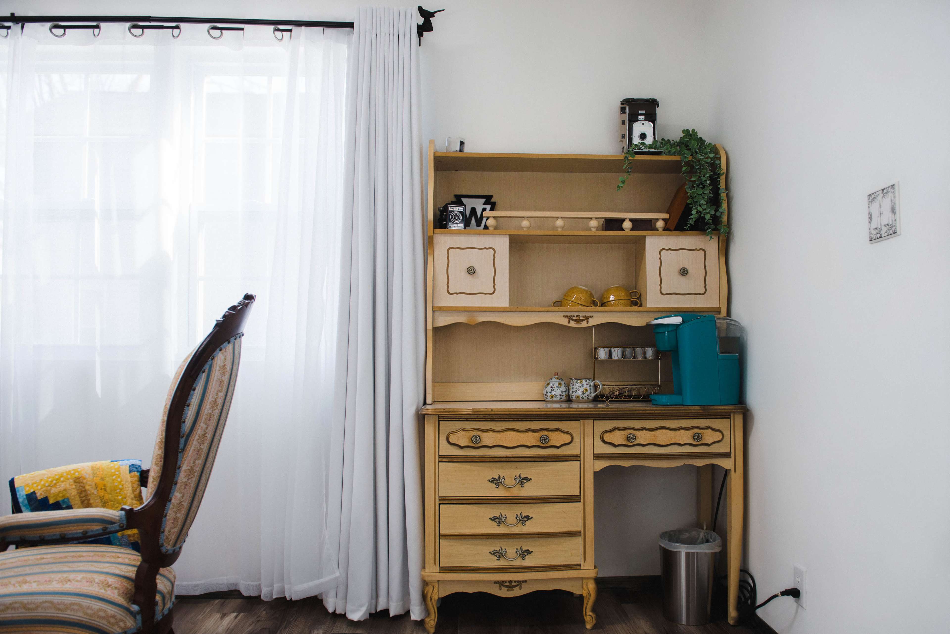 A wooden writing desk with an upper shelf, a coffee maker, and decorative items in a bright room with a white curtain and a comfortable chair.