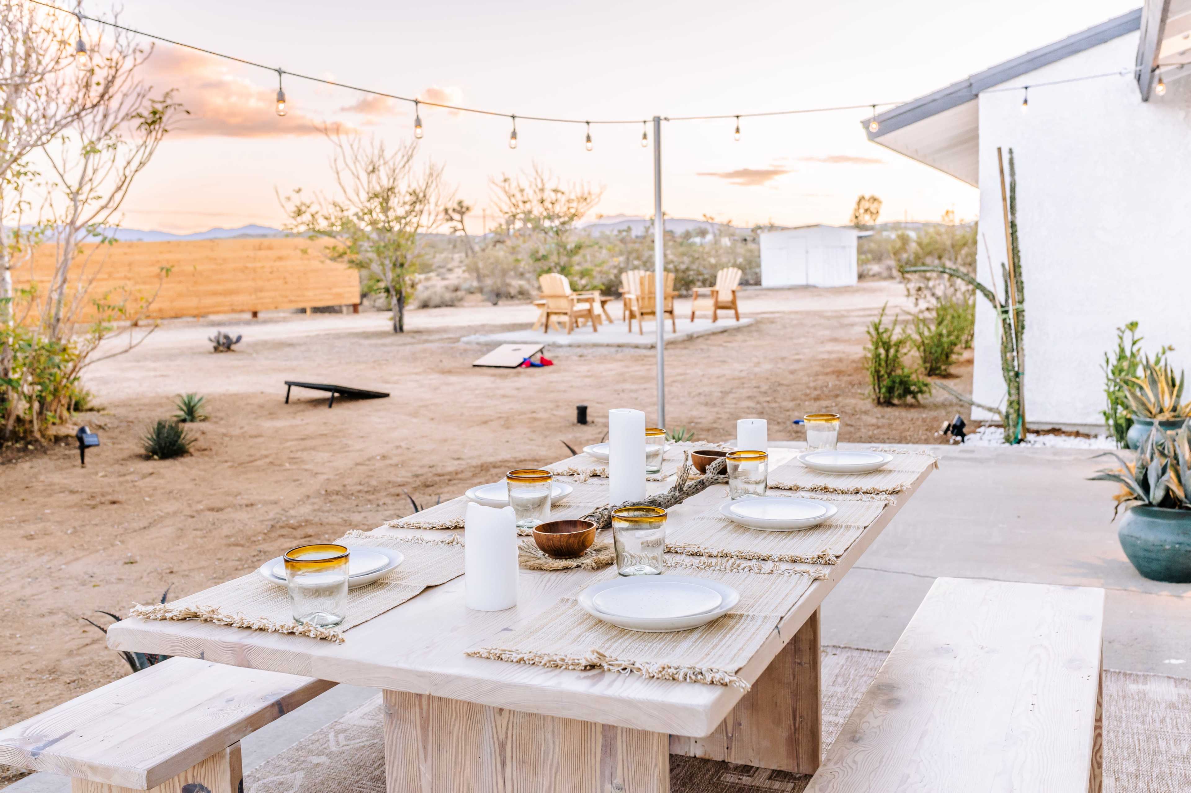 A wooden dining table is set for a meal outdoors, surrounded by desert landscaping and string lights overhead, with two chairs visible in the background.