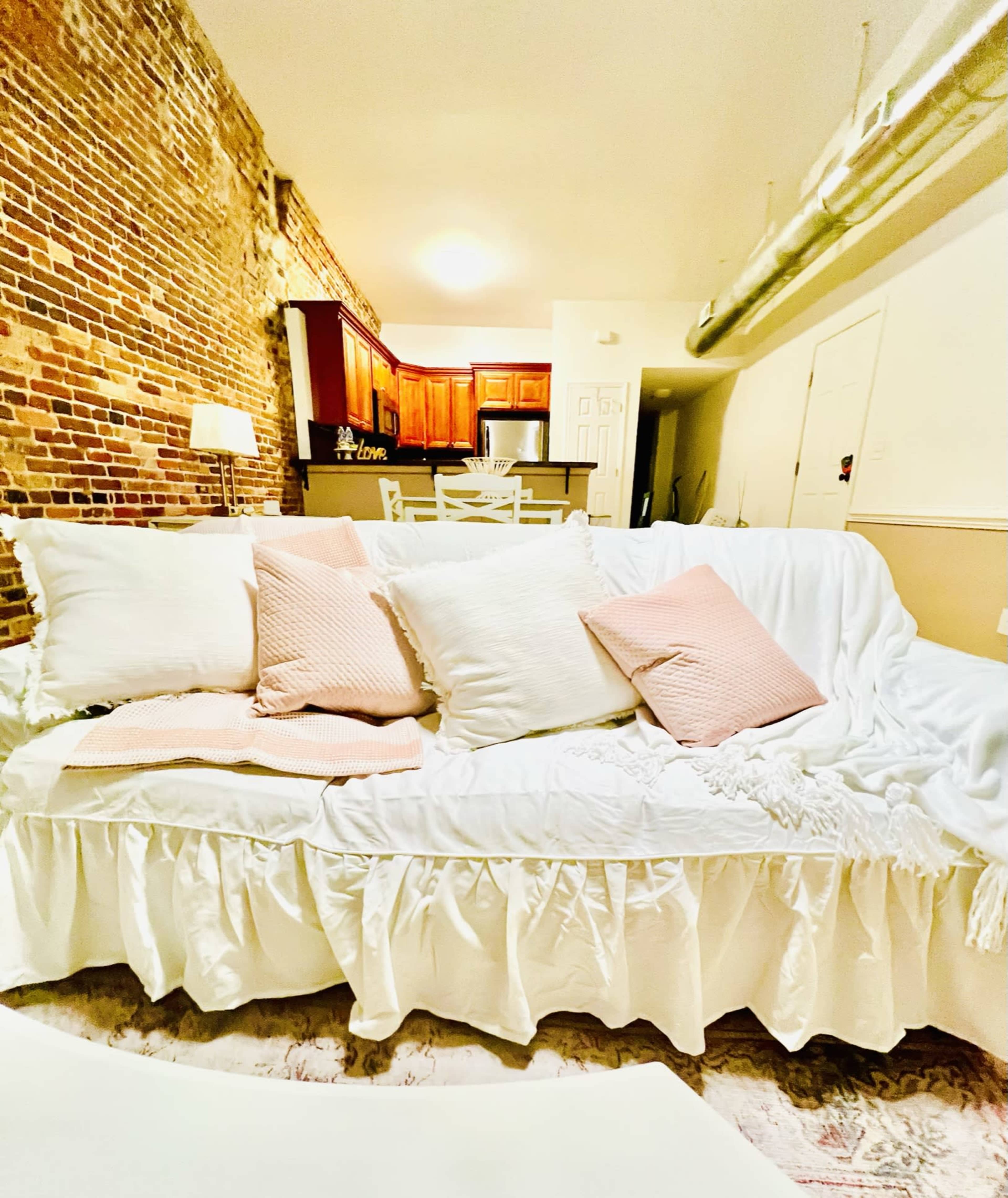 A white sofa with several pillows is positioned in a room featuring exposed brick walls and a view of a kitchen in the background.