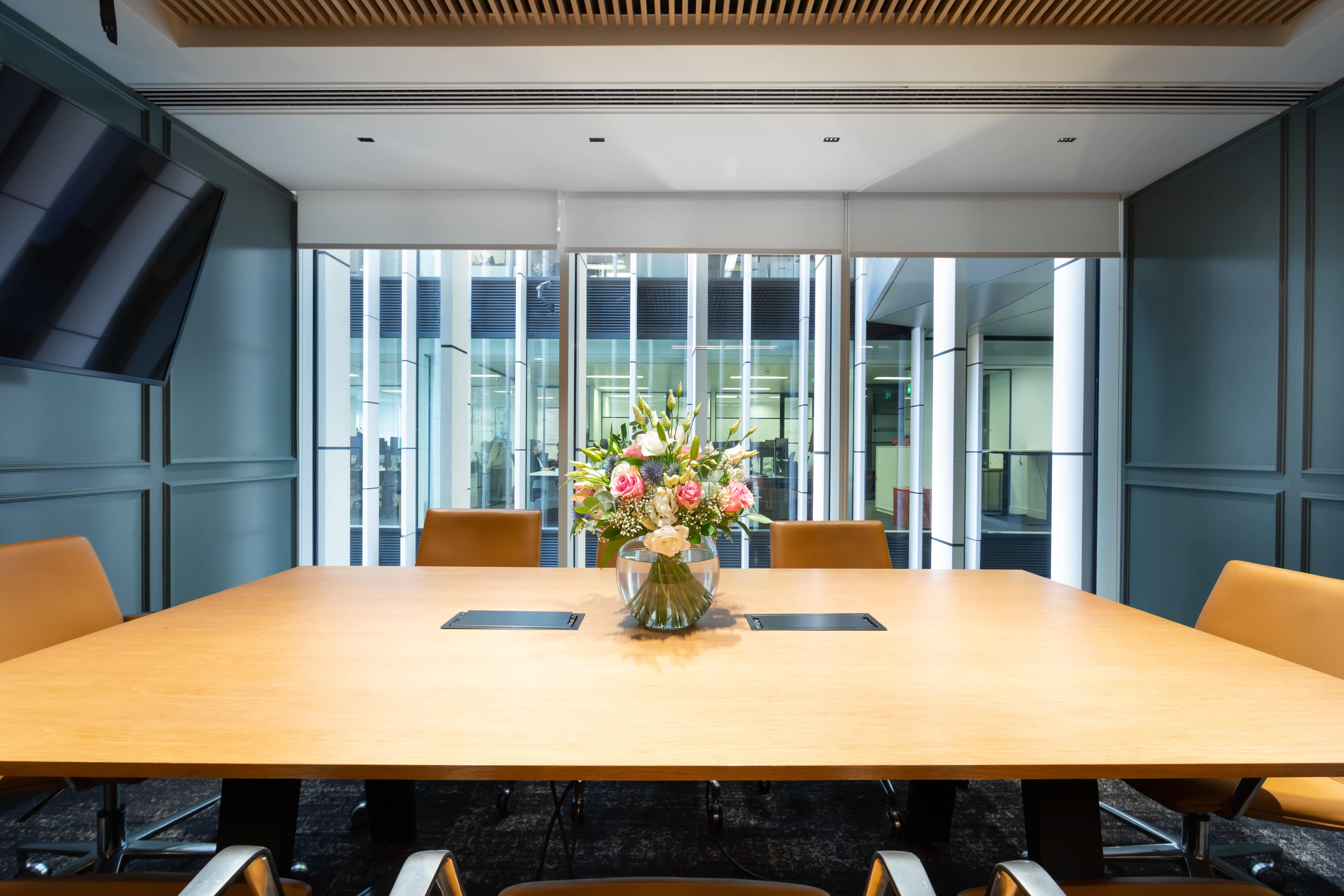 A large wooden conference table with a floral centerpiece is set in a modern meeting room with large windows overlooking a courtyard.