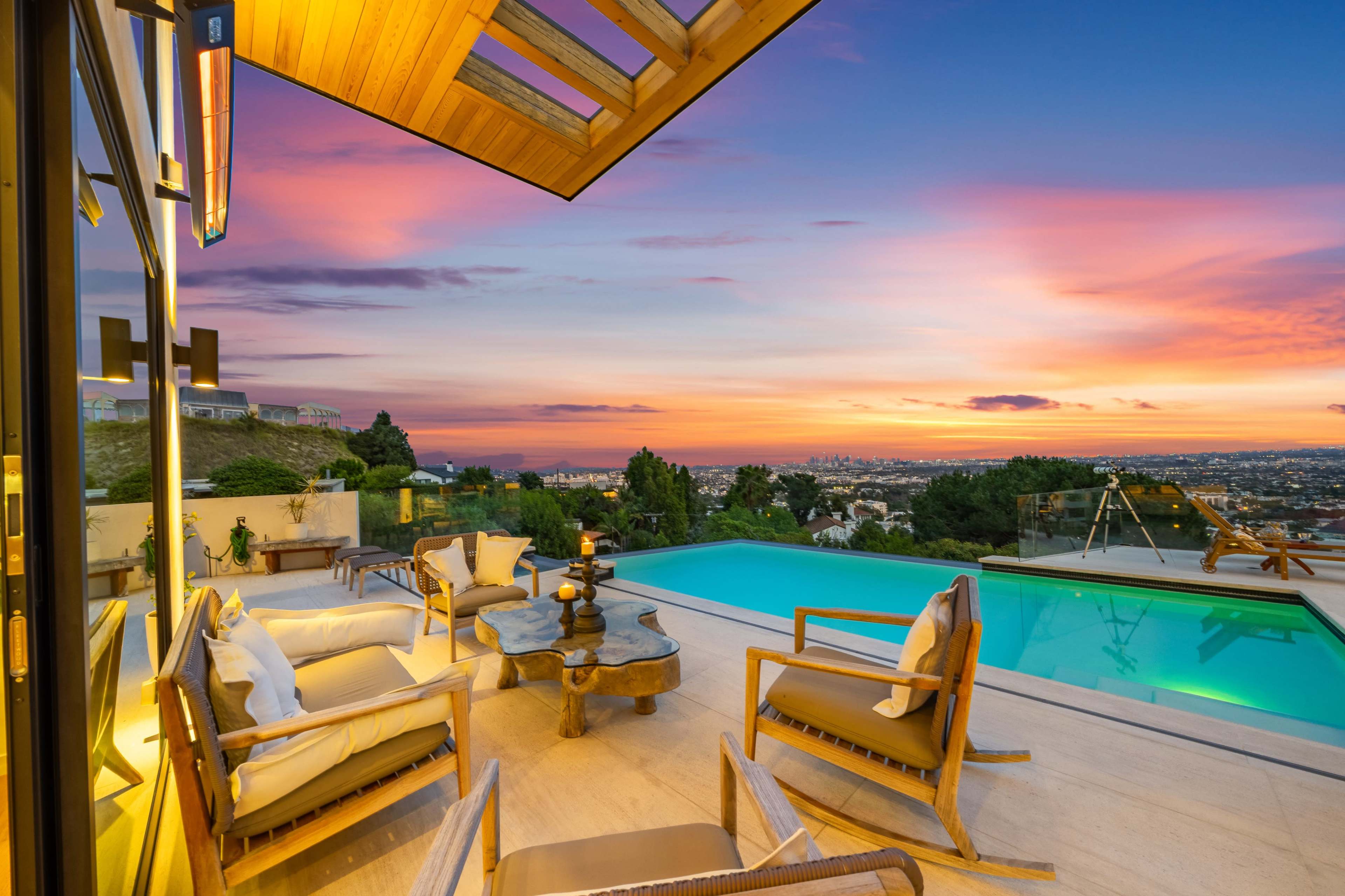 A modern outdoor patio features comfortable seating around a table, overlooking a swimming pool with a sunset view of the city skyline.