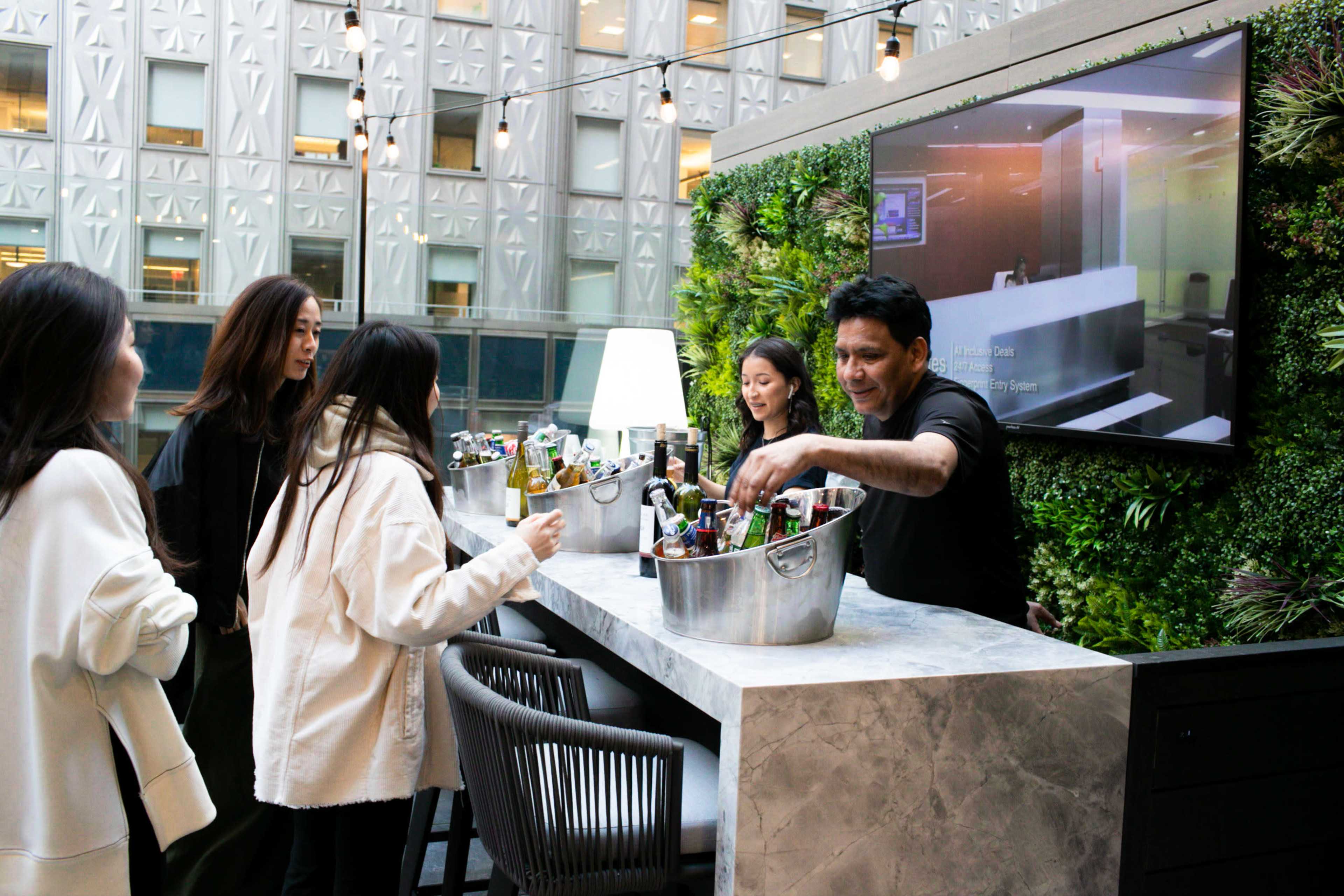 A bartender serves beverages from a bucket to a group of four women at a modern bar surrounded by greenery and a screen displaying information.