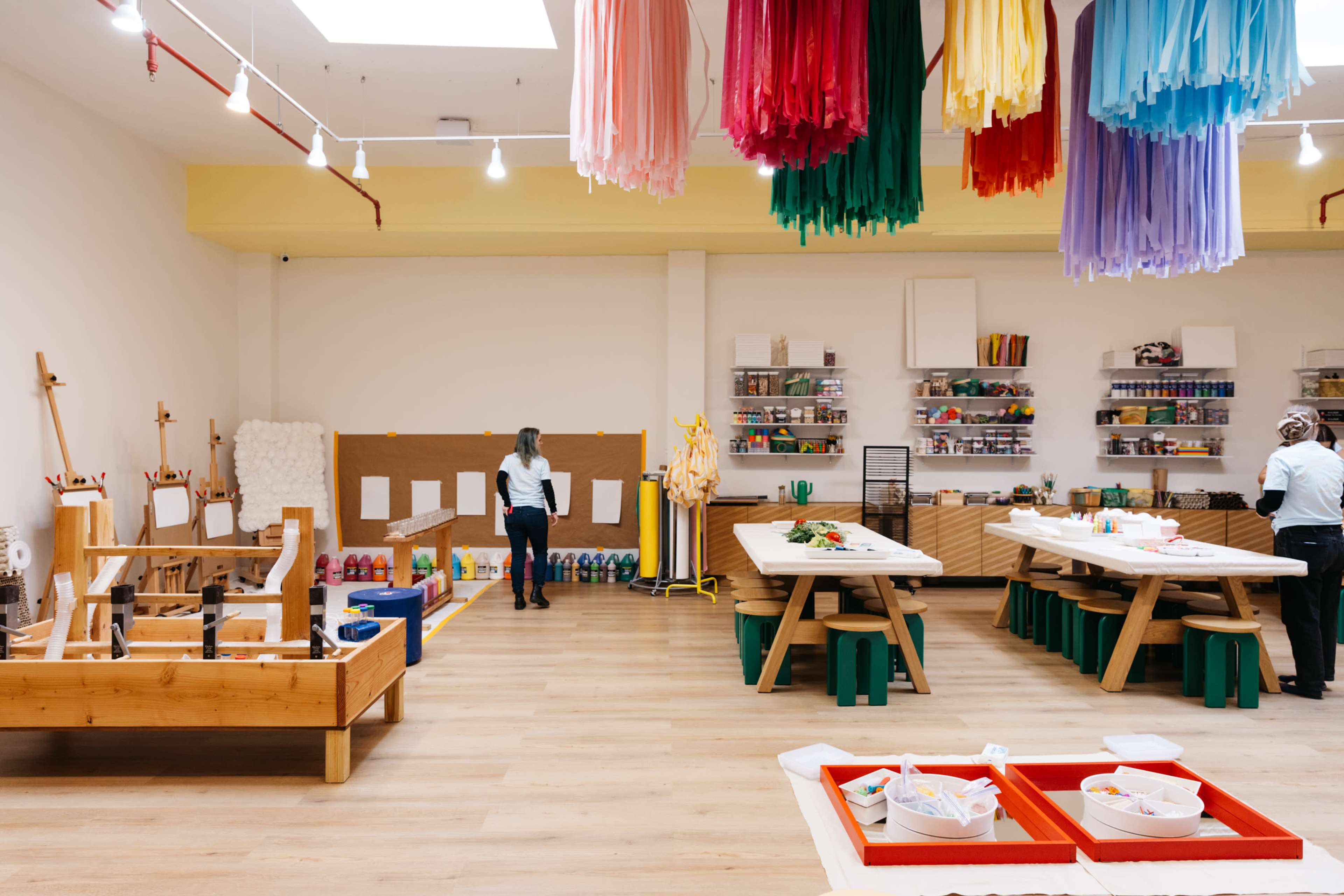 A brightly lit art studio features large wooden tables, colorful decor hanging from the ceiling, and various art supplies organized on shelves.