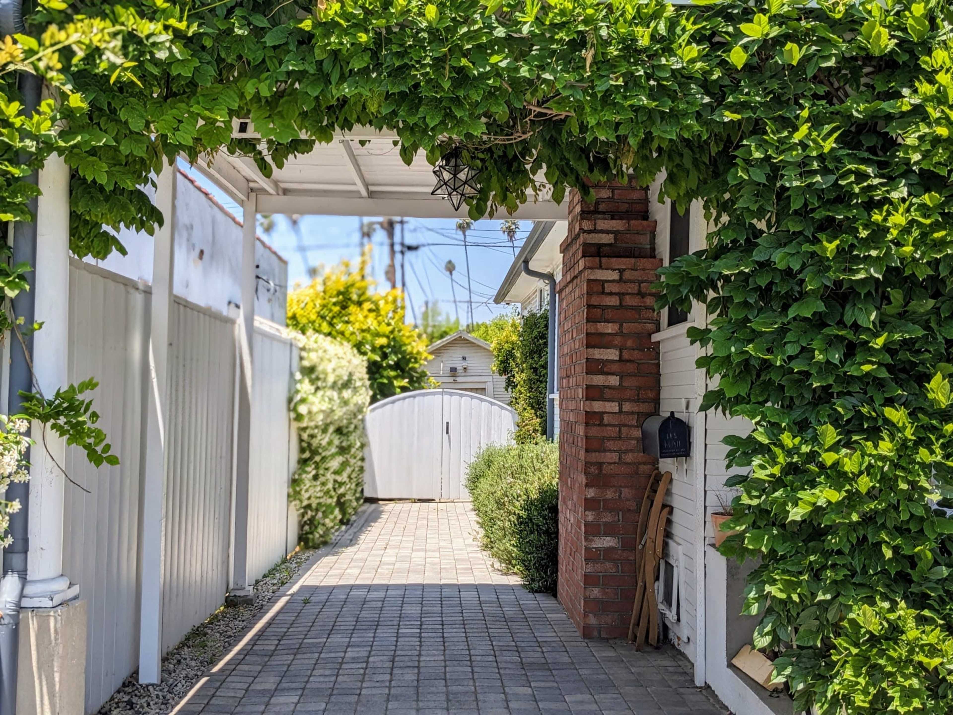 A paved pathway leads through a green archway toward a white gate at the end of a residential alley.